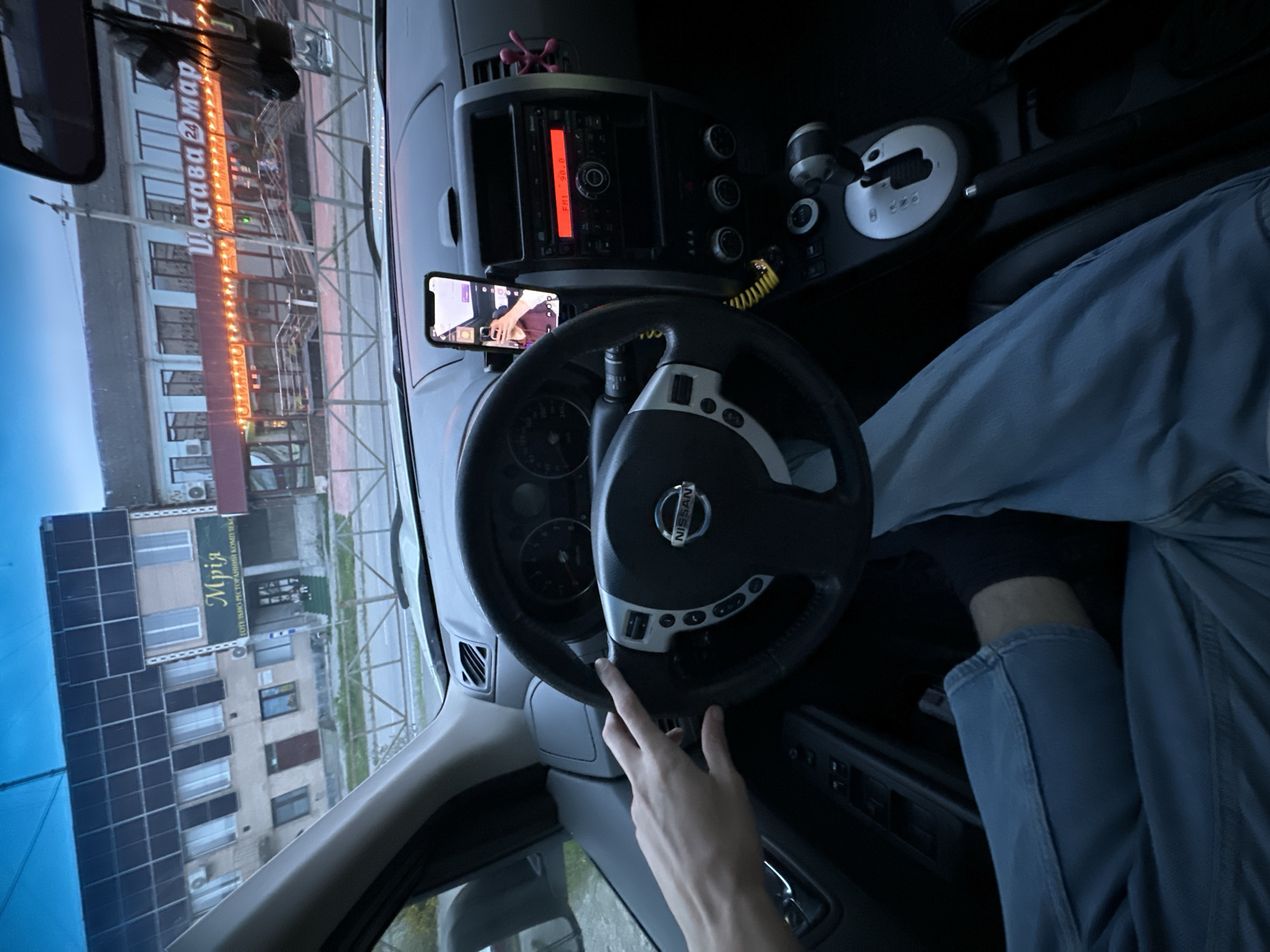 The image is taken from inside a Nissan car, showing the dashboard, steering wheel, and the driver's legs. The driver's left hand is resting on the steering wheel. A phone is mounted on the dashboard, displaying a video call with a person wearing a maroon shirt. The car is parked on a street in Shatava, Ukraine, as evidenced by the signage on buildings in the background. One building has a sign that reads "Мрія" (Dream), and another building has a sign that says "Шатава 24 Мар" (Shatava 24 Market). The sky is dark, suggesting it is either early morning or late evening. The scene is viewed from the driver's perspective.