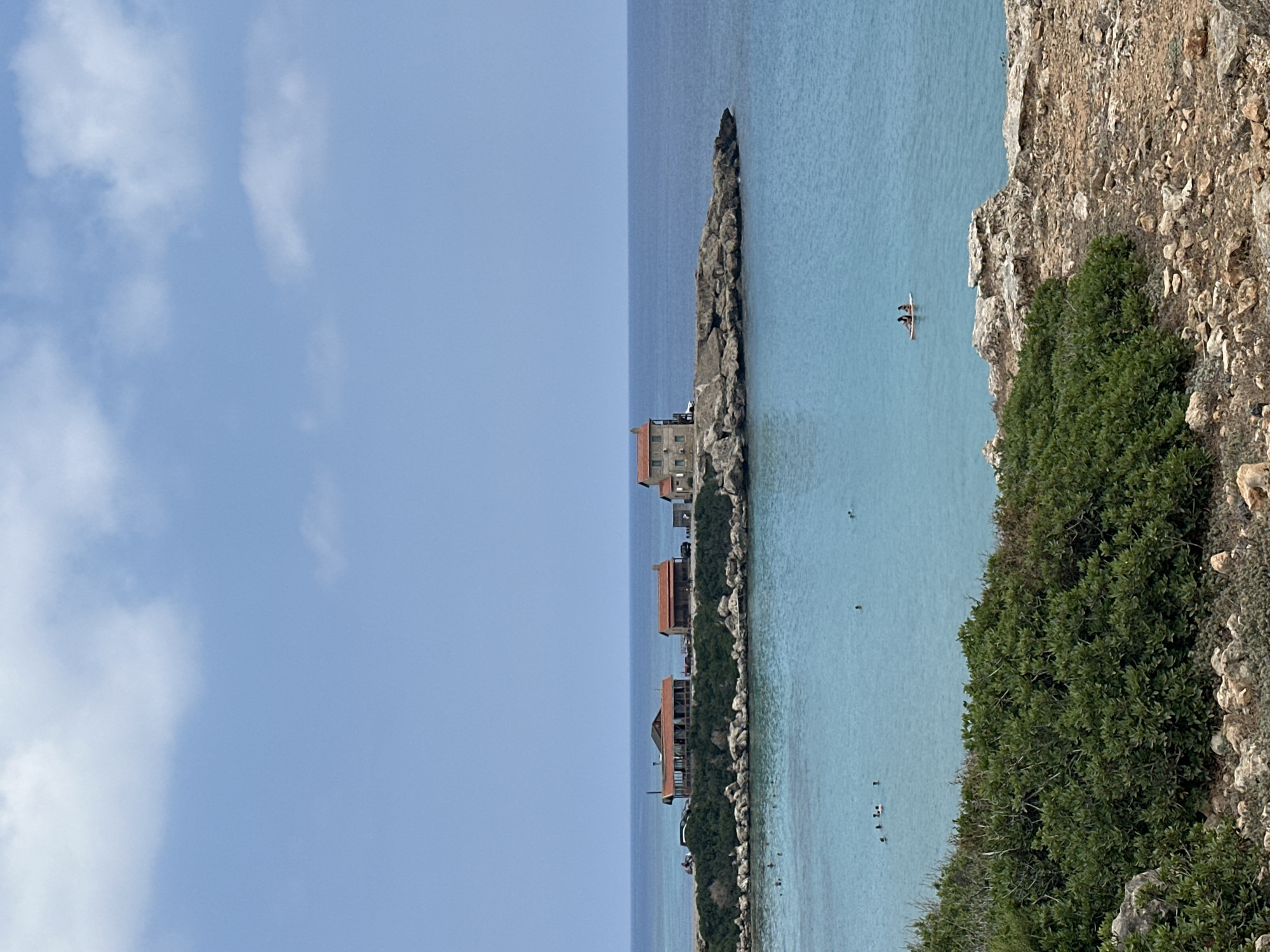 This is a landscape photograph taken from a rocky, vegetated overlook, looking out towards a calm, turquoise sea and a small peninsula. On the peninsula, there are several buildings with red tiled roofs, appearing to be houses or possibly small businesses. Some of these buildings are clustered together, while one is more isolated on the rocky edge. The sea is clear and appears shallow near the shore, with several people visible swimming or wading. Further out in the water, two individuals are on a paddleboard. The sky is a pale blue with scattered white clouds. The foreground is dominated by rough, brown rocks and dense green shrubbery. The scene suggests a coastal area, likely a beach or bay, possibly in a Mediterranean climate given the architecture and vegetation. The overall impression is one of a serene, sunny day at the coast.