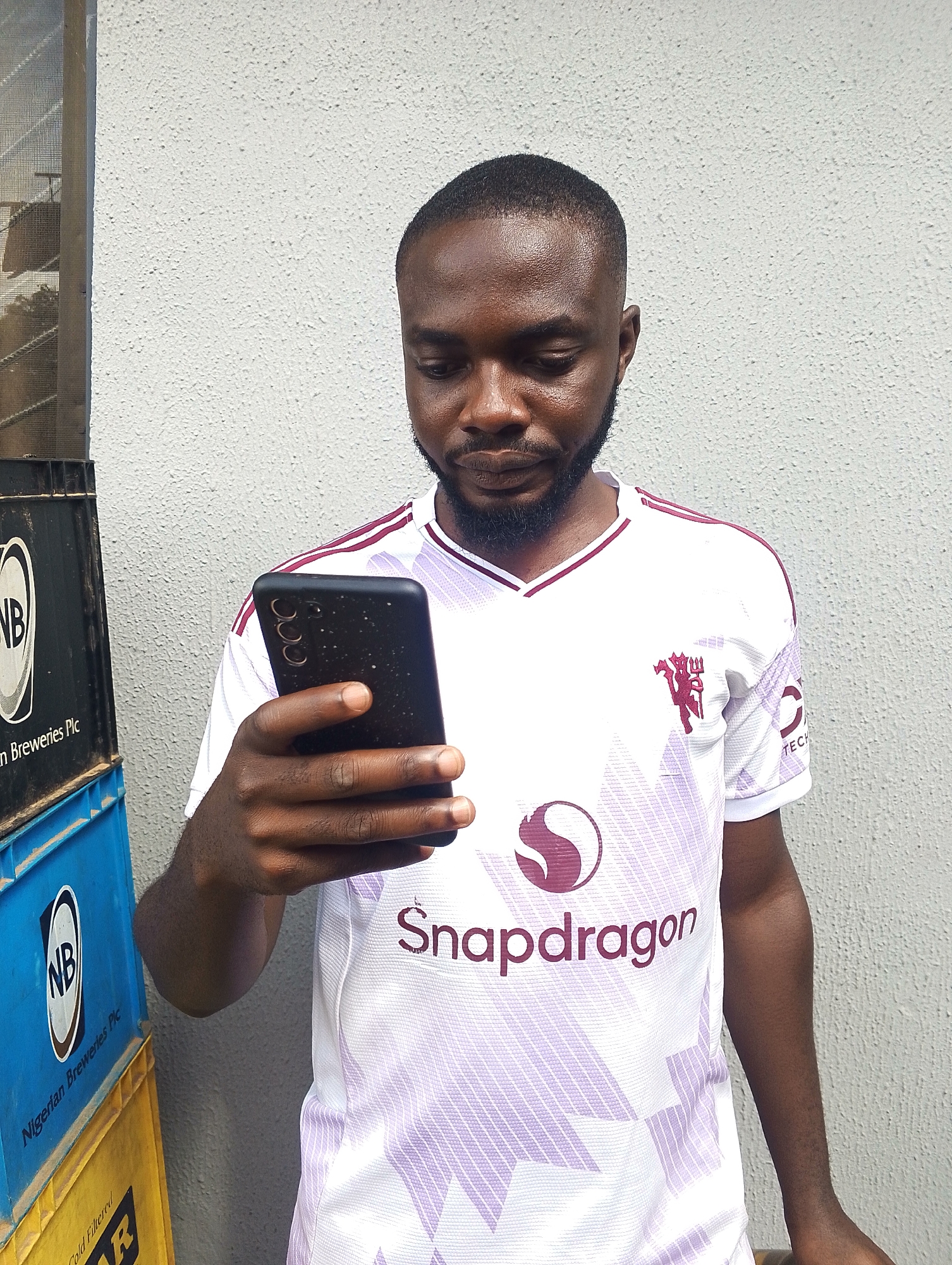 A man in a white Manchester United jersey with a Snapdragon logo is looking down at his black smartphone. He is standing in front of a textured white wall. To his left are stacked crates, one blue with the "NB" logo and "Nigerian Breweries Plc" text, and a partially visible yellow crate below it. The scene appears to be outdoors, possibly in an urban setting. The lighting suggests it is daytime.