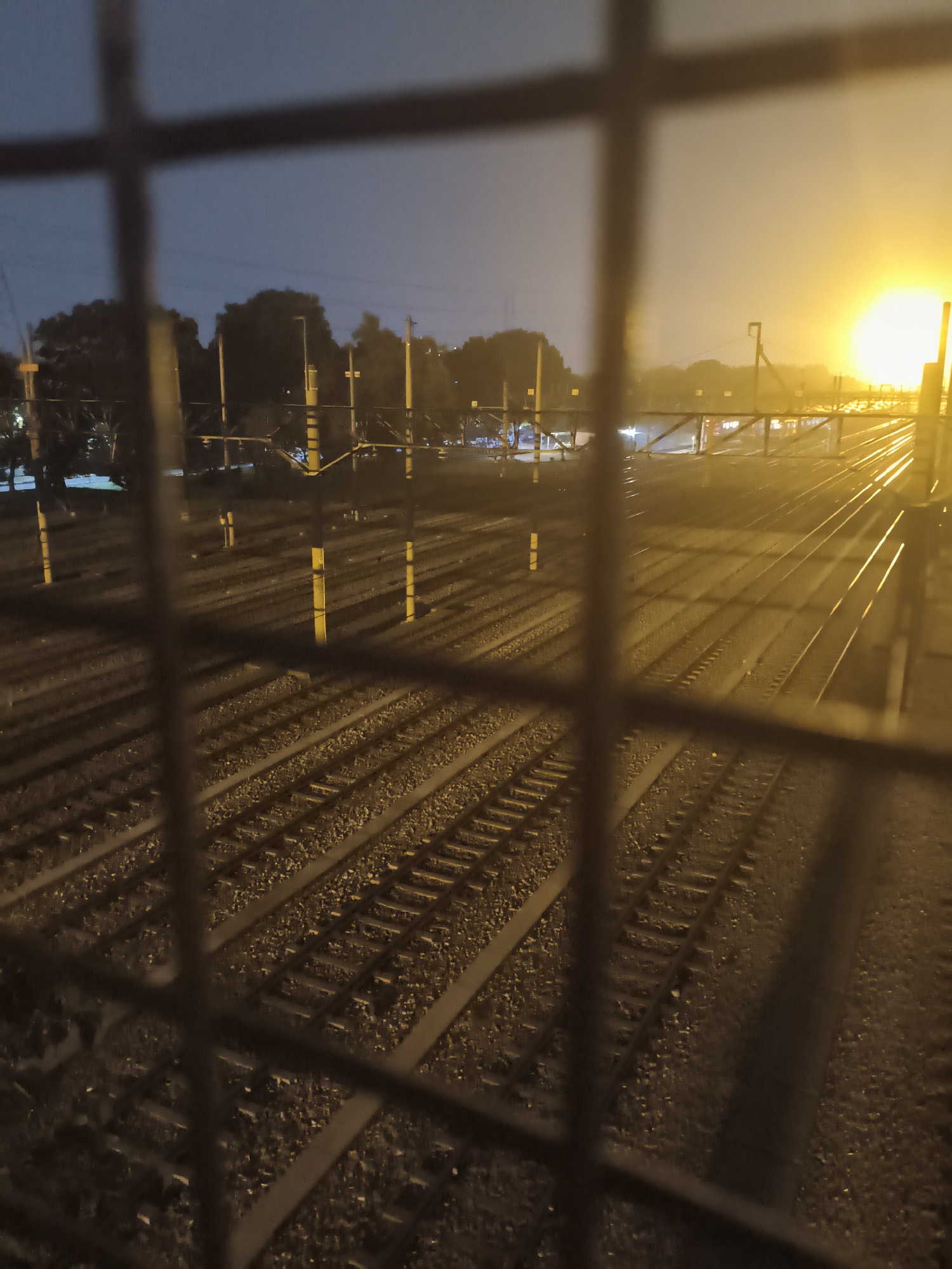 The image shows a view of train tracks at night, seen through a blurred metal grate. The sky is dark blue with a bright yellow glow in the distance, likely from a street light or a distant sun. There are multiple sets of parallel train tracks stretching into the distance, with gravel ballast between them. Several yellow poles with dark tops are visible along the tracks, possibly indicating signals or lighting. Trees are present in the background on the left side of the image, and a structure that appears to be a pedestrian overpass or bridge crosses the tracks further in the distance. The overall lighting suggests it is either dawn, dusk, or a heavily lit night scene. The location is identified as Depok, Indonesia.