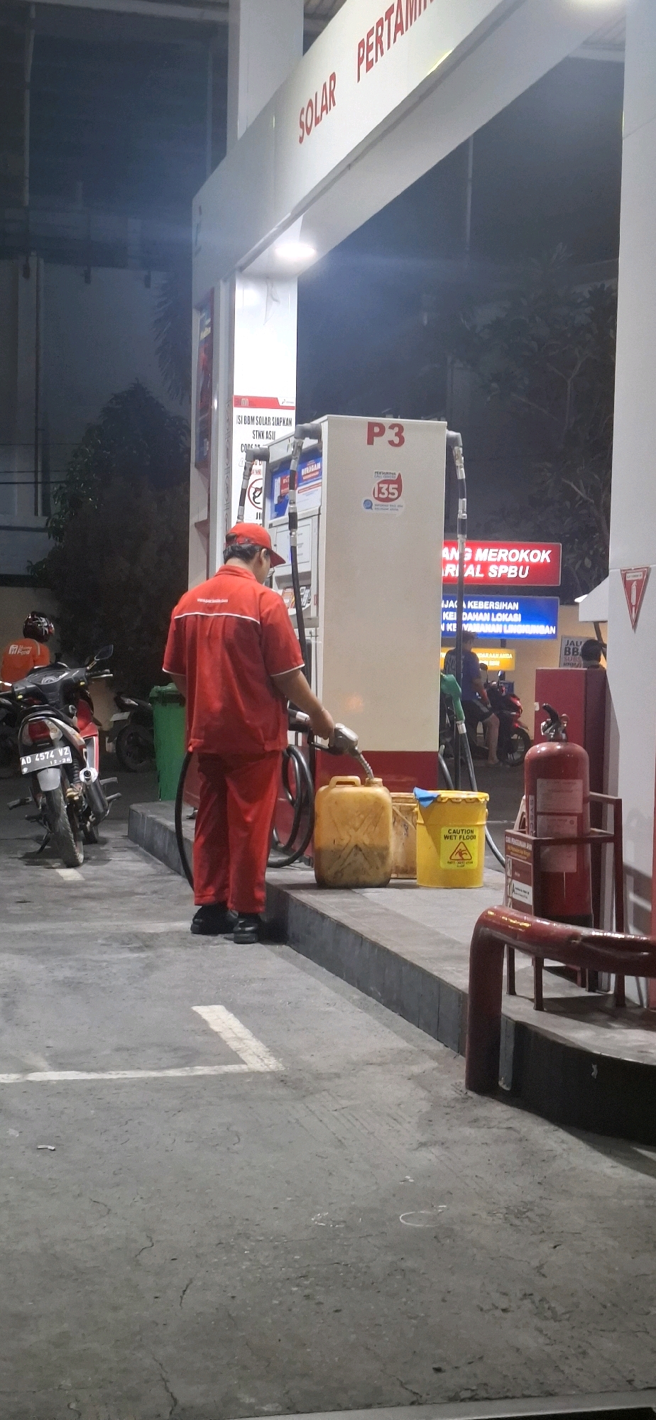 At a Pertamina gas station in Colomadu, Indonesia, a gas station attendant in a red uniform and cap is dispensing fuel from a pump into a yellow plastic jerrycan. The attendant is standing near a gas pump labeled "P3". In the background, motorcycles are parked, and a "DILARANG MEROKOK" (No Smoking) sign is visible. A yellow bucket with a "CAUTION WET FLOOR" sign is also present near the jerrycan. The scene takes place at night, with artificial lighting illuminating the area.