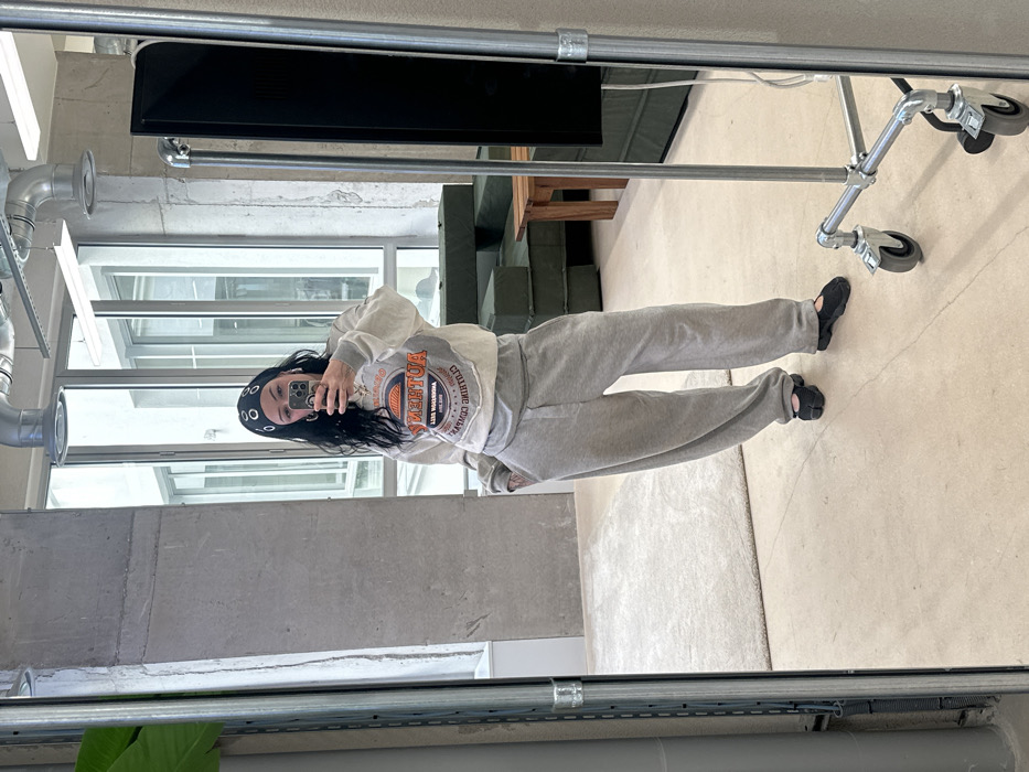 A full-length mirror selfie depicts a female subject standing in a modern, industrial-style interior in Paris, France. The subject has dark, wavy hair, a black headband with metal eyelets, hoop earrings, and visible tattoos on her left forearm. She is dressed in a light grey cropped sweatshirt featuring an orange and blue "AUTHENTIC SPORTWEAR" graphic, oversized light grey sweatpants, and black tabi-style flat shoes. She holds a smartphone with a dark case, angled to capture her reflection. The background, visible in the mirror, features exposed concrete walls, a light-colored floor, and a beige shaggy rug. Large windows reveal an exterior modern building. Overhead, silver industrial ductwork and linear strip lighting are present. Furniture includes a dark green modular sofa and a light wooden coffee table. A metal rolling rack with a dark screen or panel stands on the right, and a tall green plant is partially visible on the far left.