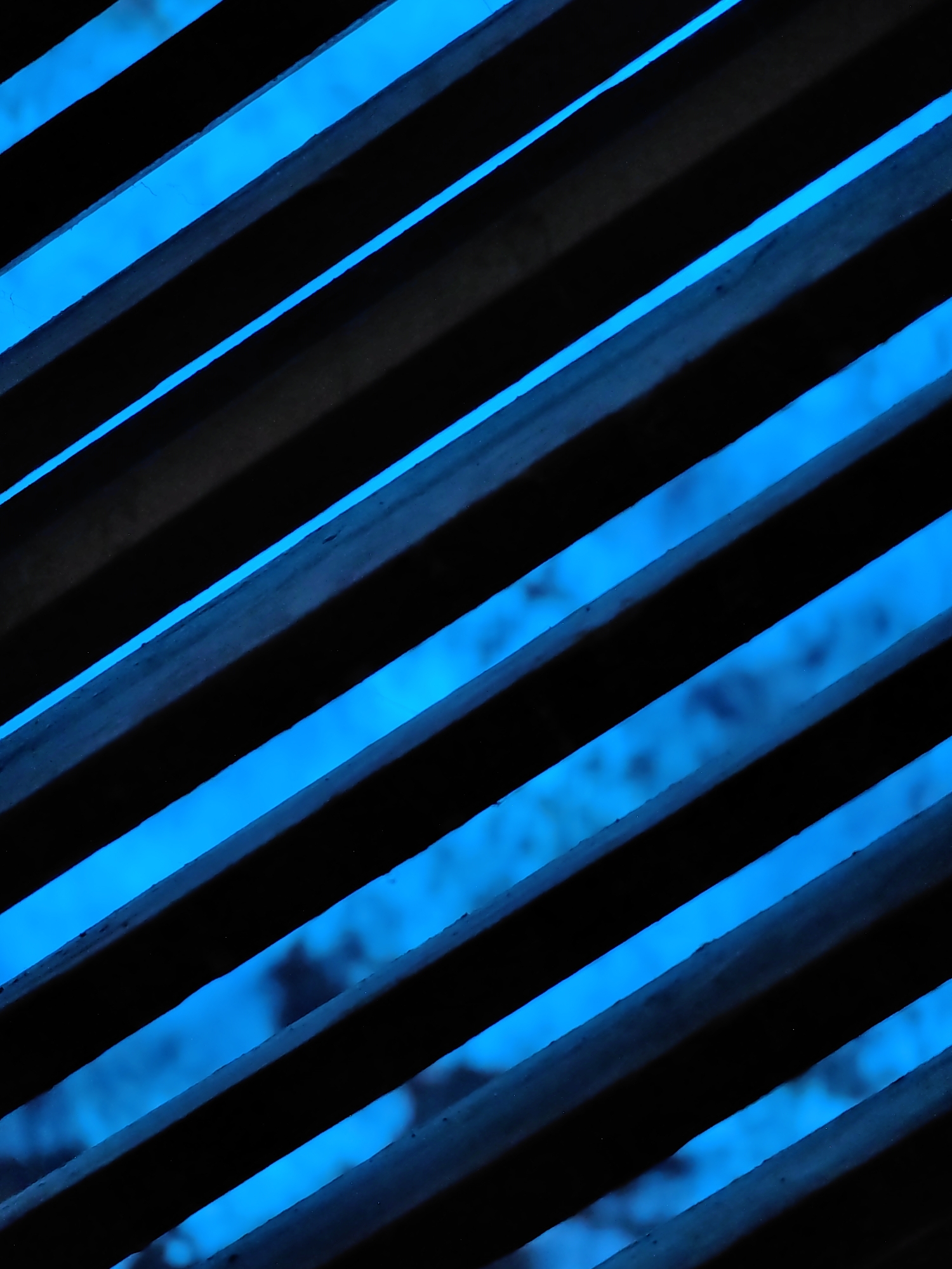 This is a close-up, abstract photograph of dark, diagonal slats with bright blue light filtering through the gaps. The perspective is from below, looking up through what appears to be a grille or shutter system. The blue light illuminates the spaces between the slats, creating a stark contrast with the dark, solid bars. The background visible through the slats is out of focus and appears to be a mottled blue-green, suggesting foliage or a distant, dimly lit environment. There are no people or specific objects clearly discernible. The scene is devoid of any distinguishing features that would allow for specific location identification within Vashisht, India, or any other city. The lighting and composition suggest an outdoor setting, possibly at dusk or night, or an interior space with a strong external light source.
