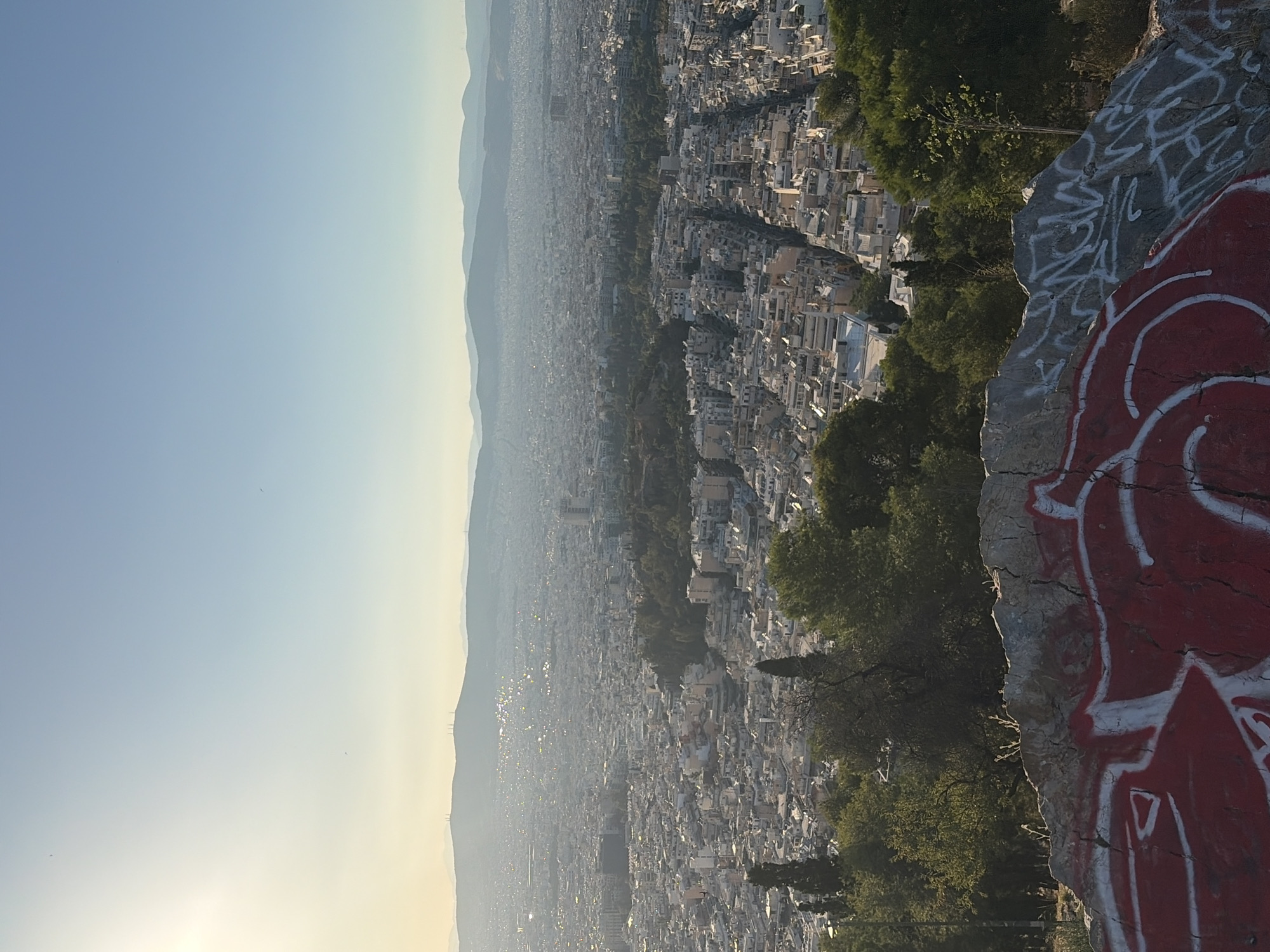 A high-angle, full outdoor, eye-level shot shows a sprawling city with a bright sky and distant mountains. The foreground features a rough, grey rock face covered in red and white graffiti. Beyond the rock, the middle ground is filled with densely packed, low-rise buildings in shades of white and beige. Several patches of dark green trees are interspersed throughout the urban landscape. In the distance, the city continues to spread out, eventually meeting a hazy line of blue-grey mountains under a pale blue sky. The sun is visible in the upper left corner, casting a bright glow. Several faint, dark specks, possibly birds or insects, are scattered across the sky. The overall impression is of an expansive urban environment bathed in natural light.