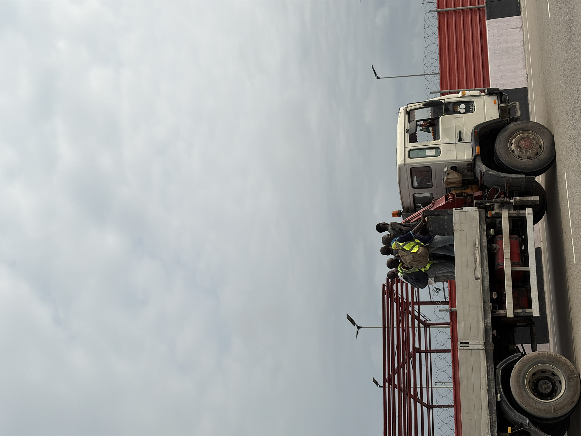 In Igboefon, Nigeria, a flatbed truck with a crane attachment is captured in motion. The truck's cargo bed is occupied by a group of approximately seven Black males. Some of the men are wearing yellow high-visibility vests. The truck is traveling on a paved road, with a red barricade and barbed wire fencing visible in the background, suggesting the presence of a construction site or a restricted area. The sky is overcast with a dense cloud cover.