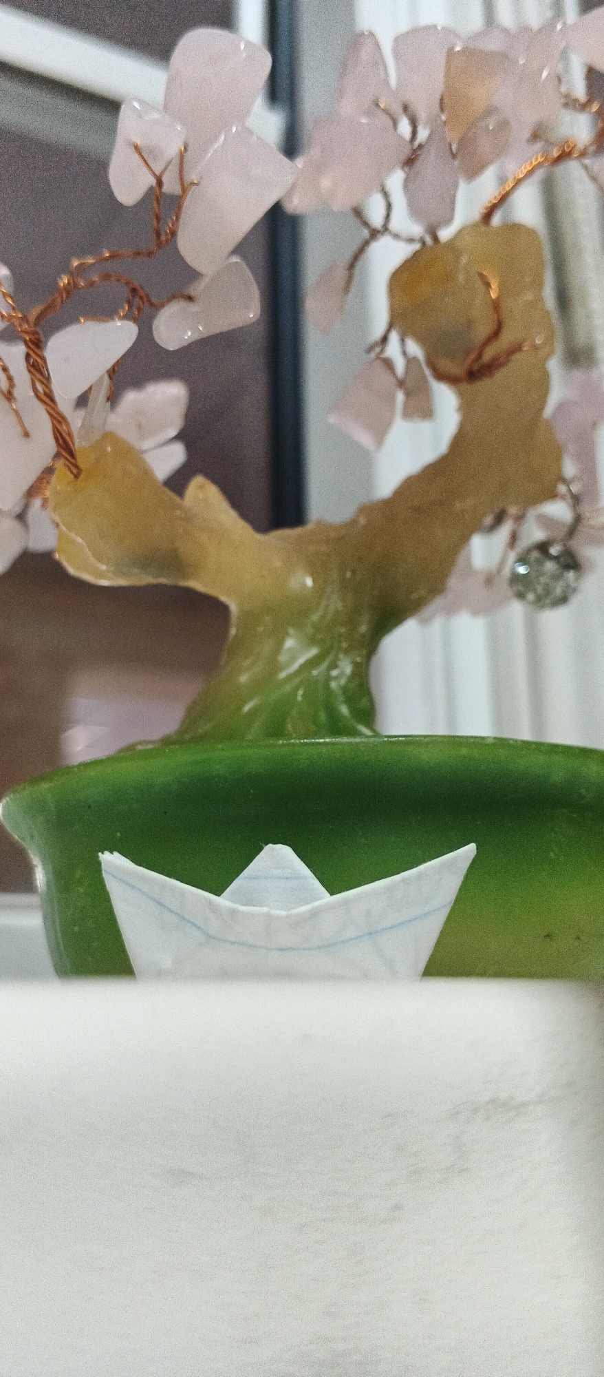 A close-up, vertical, eye-level shot shows a small, white origami paper boat positioned in the foreground. The boat, with blue lines visible, rests on a light-colored surface. Behind the boat, a glossy, deep green ceramic planter is visible. Emerging from the planter is a decorative tree, its trunk a translucent, yellowish-green, with a gradient effect. The branches of the tree are made of thin copper wire, adorned with light pink and yellowish-orange crystalline "leaves" and a small, sparkling embellishment on the right side. The background is blurred, showing hints of a window frame and vertical blinds. The scene is indoors, with soft lighting that creates some reflections on the glossy surfaces. The image does not provide any information about its location in Smila, Ukraine.