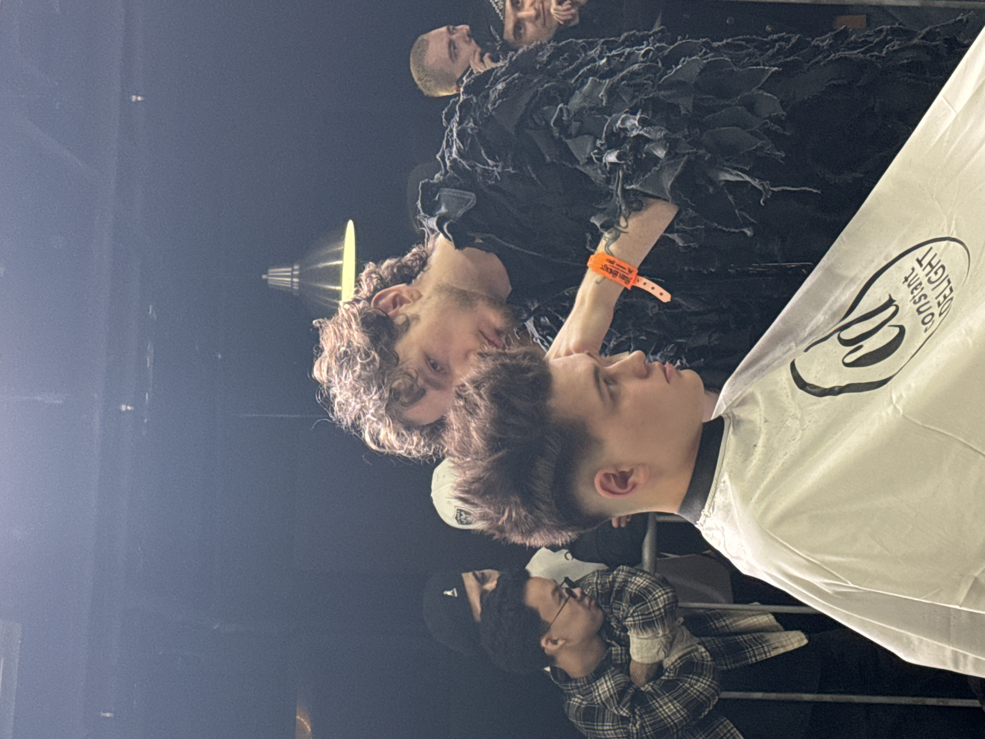 The image depicts a group of people at what appears to be a concert or event in Unknown, Unknown. A man with curly hair wearing a dark, shredded jacket has an orange wristband. A person in a white cape with "Constant Delight" logo is also visible. Other people are blurred in the background. The environment is dark with a spotlight shining.