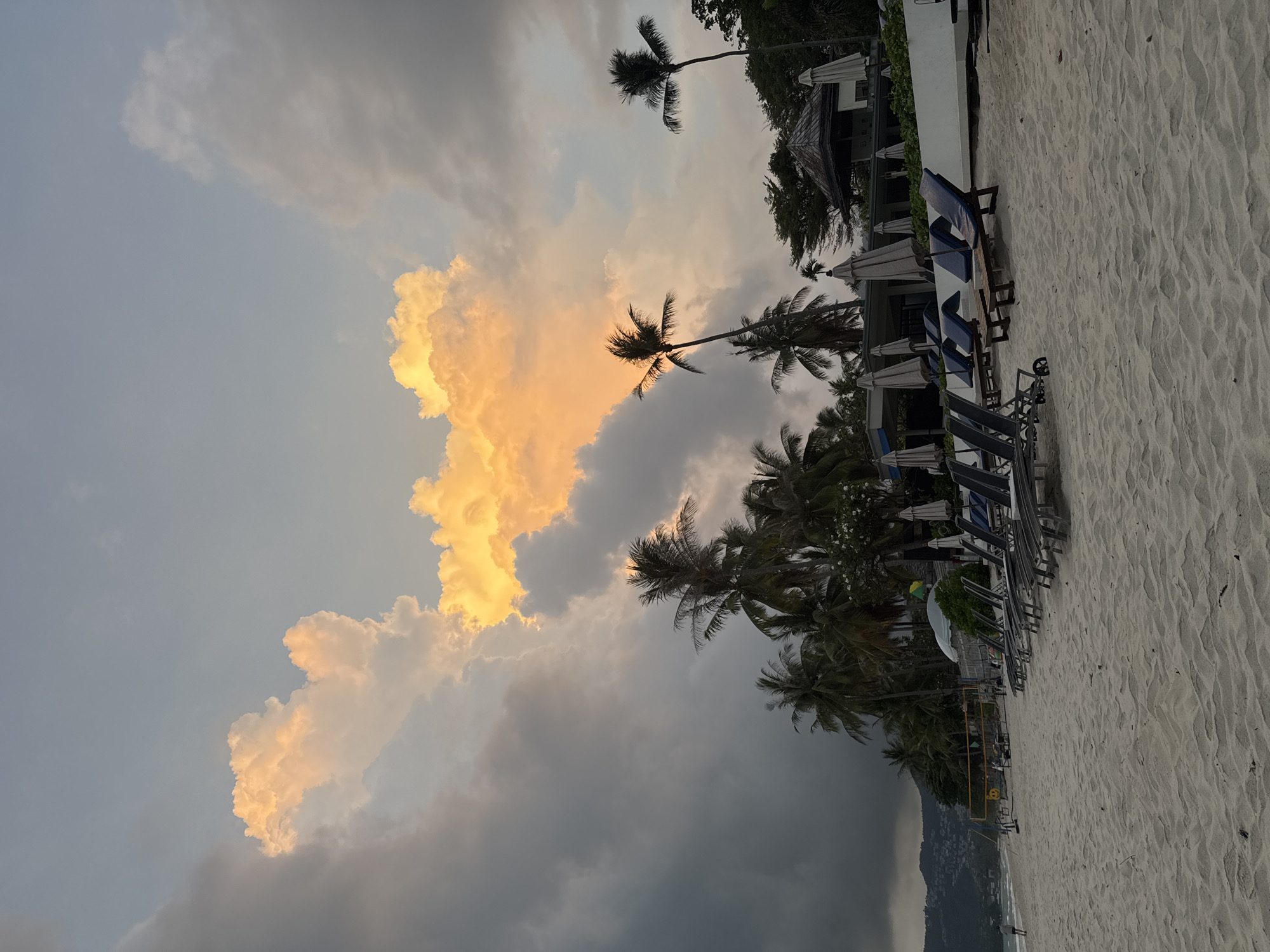 The image shows a beach scene in Baan Chaweng, Thailand, at dusk. The beach is sandy and lined with lounge chairs and umbrellas. Palm trees are interspersed along the beach. In the background, buildings and lush greenery are visible. The sky is dominated by dramatic clouds, some dark and ominous, while others are illuminated with a golden glow, likely from the setting sun. The overall impression is a peaceful tropical scene with a dynamic and somewhat ominous sky. A person is walking along the water's edge.