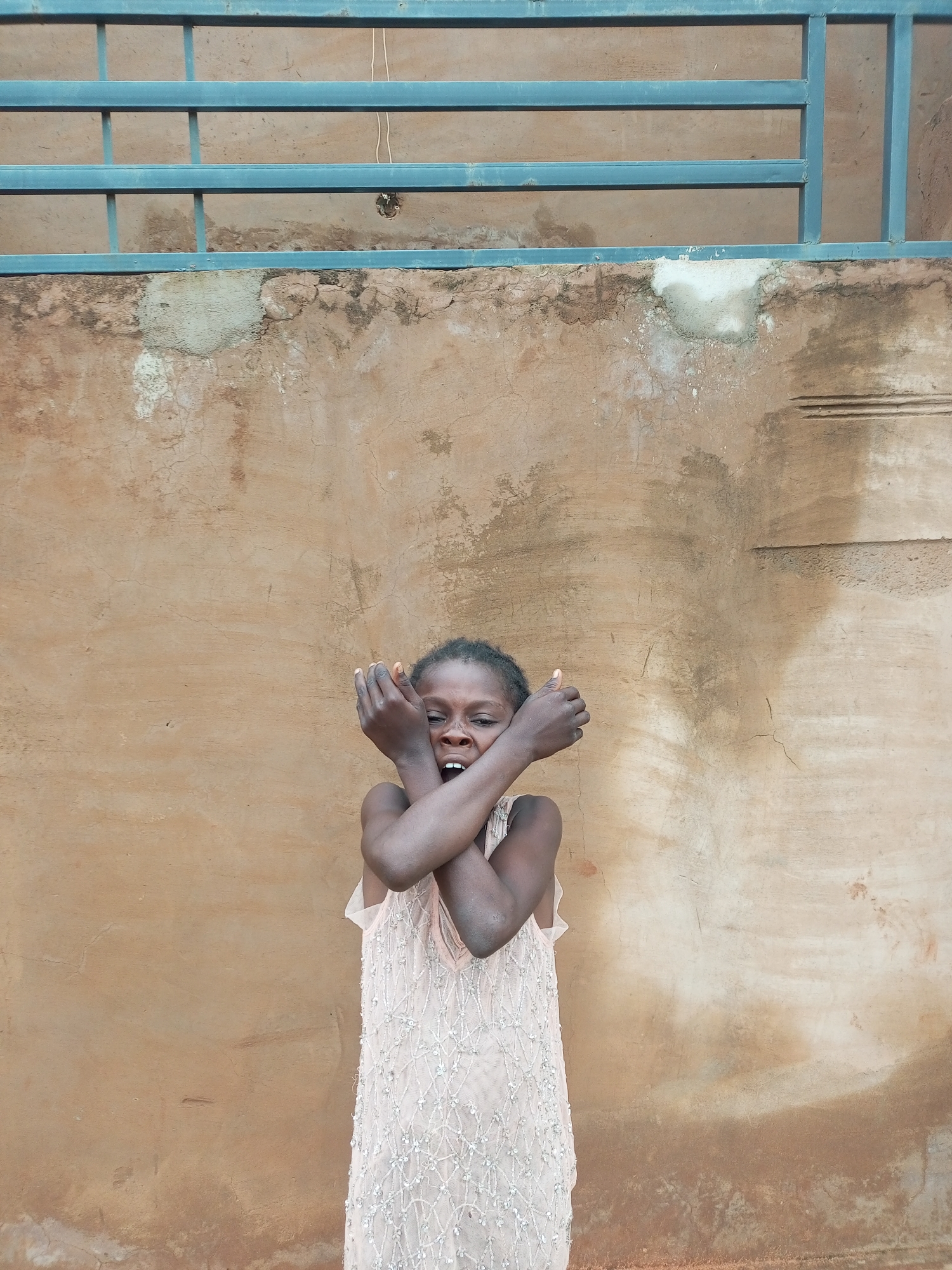 A young Black girl stands in front of a textured, ochre-colored wall, her arms crossed in front of her chest, hands clasped over her mouth. She wears a light pink, sleeveless dress adorned with small, clear beads in a diamond pattern. The wall behind her is weathered and shows signs of repair or patches of lighter material. Above the wall, a blue metal railing is visible, supported by vertical posts. The girl's expression is one of distress, with her mouth open and eyes wide. The scene is set outdoors, with the natural light casting shadows on the wall. There are no other people or objects clearly visible in the frame that provide further context for the location within Tempure, Nigeria.
