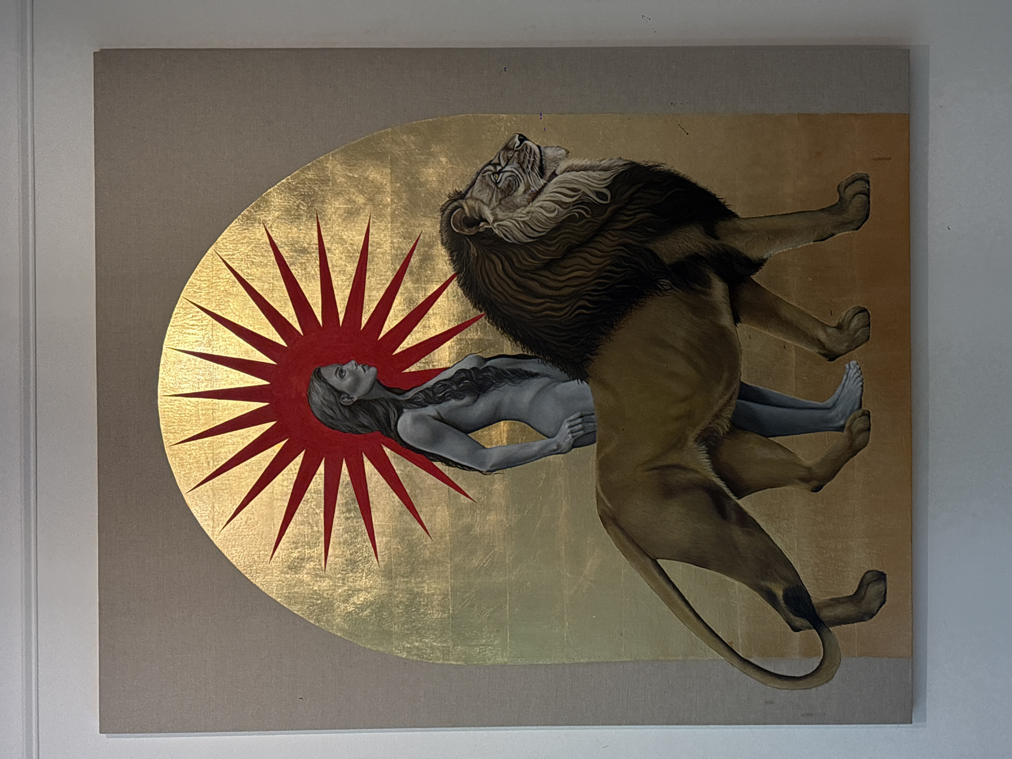 A painting is displayed on a wall in a building in Mexico City, Mexico. The painting depicts a large lion dominating a human figure. The lion, with its mouth open as if roaring, is positioned above the human figure. The human figure is oriented horizontally and seems to be held or supported by the lion. Behind the human figure, there is a radiant burst design, with a gold leaf background and red radial lines emanating from behind her. The rest of the background is a beige color. The painting is rectangular and mounted on a wall in what appears to be an interior setting.