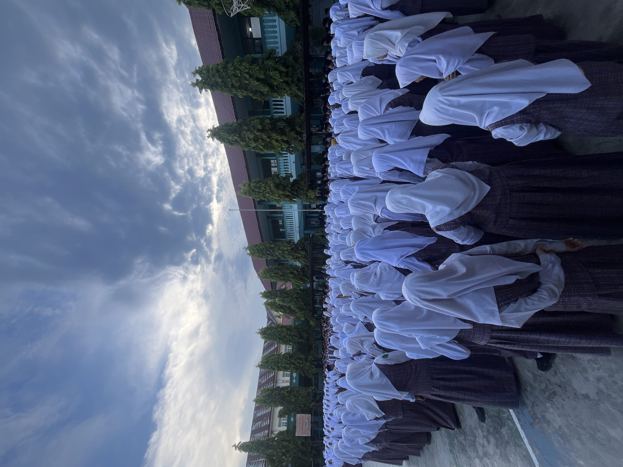 A large group of uniformed students, predominantly female and wearing white hijabs and plaid skirts, stand in formation on a concrete courtyard. Behind them, a multi-story building with green accents and a red tiled roof is visible, suggesting a school or educational institution. Tall, dark green trees are planted in front of the building, and the sky above is partly cloudy with a mix of gray and lighter tones, indicating either early morning or late afternoon. The overall scene captures a moment of organized assembly, possibly a flag ceremony or morning assembly, in Lhokseumawe, Indonesia.