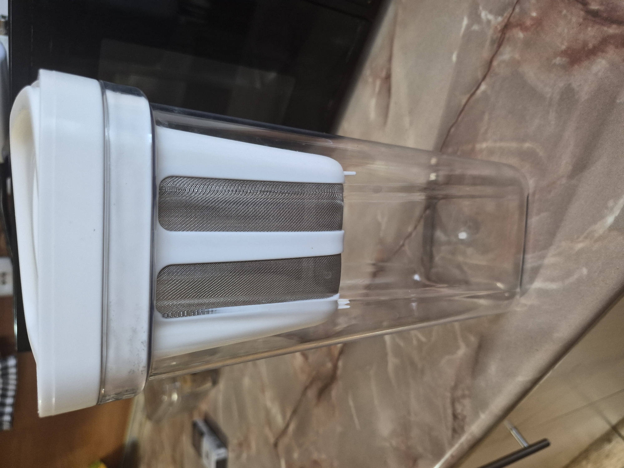 A rectangular, clear plastic pitcher or container is prominently displayed, lying on its side. It features a white, flat lid on one end and an internal, cylindrical filter assembly, also white, with fine metallic mesh on its sides. The mesh appears silver or dark gray. The clear plastic body of the container shows some reflections from the surrounding environment.
The container rests on a speckled or marbled surface, exhibiting shades of beige, brown, and hints of red, likely a kitchen countertop. In the background, dark, blurred areas suggest other kitchen elements or appliances. A dark, possibly black, surface is visible behind the container, and a silver drawer pull or handle can be faintly seen in the lower right corner, further indicating an indoor, possibly kitchen, setting.
No people are present in the image. No specific activity is taking place; the object is static. The time of day and weather are not discernible from the indoor lighting. There is no visible text on the object or in the scene. The context provided specifies the location as Yerevan, Armenia.