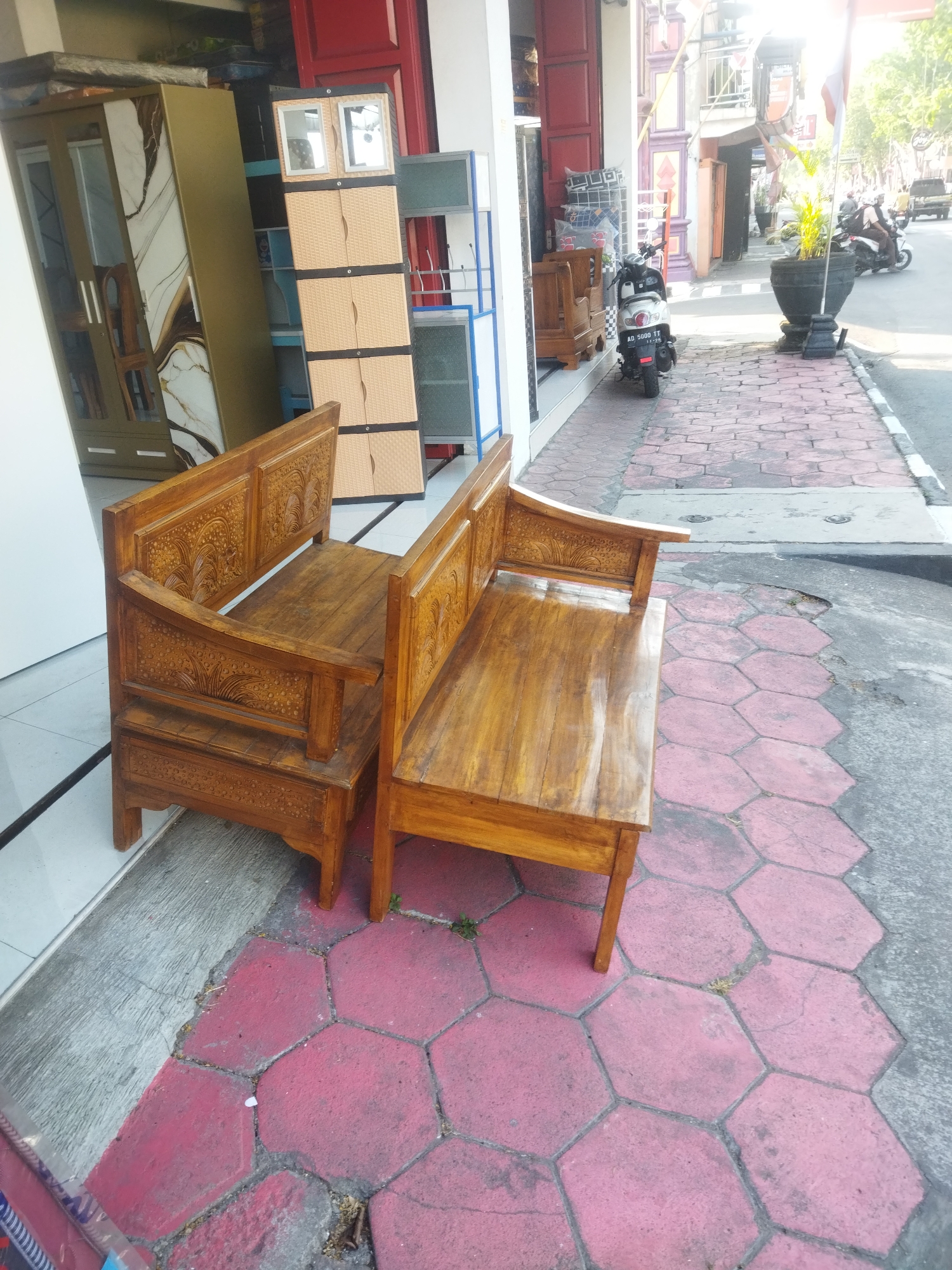 This media file shows a street scene in Sukoharjo, Indonesia. In the foreground, two intricately carved wooden benches are placed outside what appears to be a furniture store. The benches are made of dark wood and feature detailed floral and leaf patterns on the backrests and armrests. Behind the benches, inside the store, a tall, multi-compartment plastic cabinet is visible, along with other furniture items including a wardrobe with a marbled design.

The scene extends to the sidewalk and street. A motorbike with license plate AD 5080 TT is parked on the sidewalk. Further down the street, several people on motorbikes are visible, suggesting a typical day with traffic. Large potted plants flank the sidewalk, and an Indonesian flag is displayed. The lighting suggests it is daytime, likely mid-morning or afternoon, with bright sunlight casting shadows. The weather appears to be clear. The hexagonal pavement tiles on the sidewalk are a notable detail, with some showing wear and tear. The overall impression is that of a bustling street in a commercial area of Indonesia.
