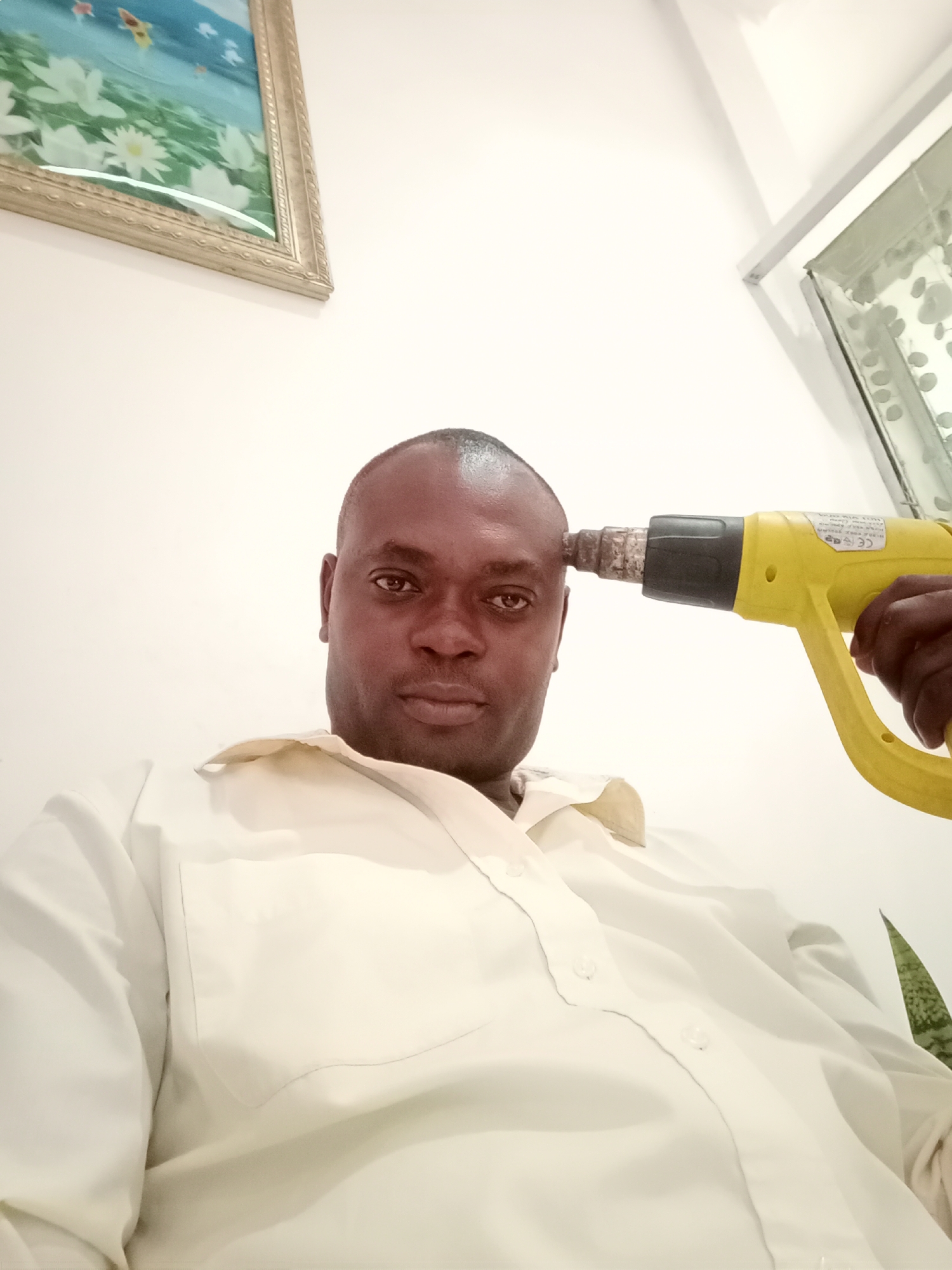 The image depicts a dark-skinned man with short, dark hair. He is wearing a light-colored, button-down shirt. A yellow heat gun is positioned to touch his forehead. The background is a plain white wall with a framed painting in the upper left corner and another framed object or window in the upper right corner. The setting is likely indoors, with the location potentially being Gwarinpa, Nigeria, based on given information.