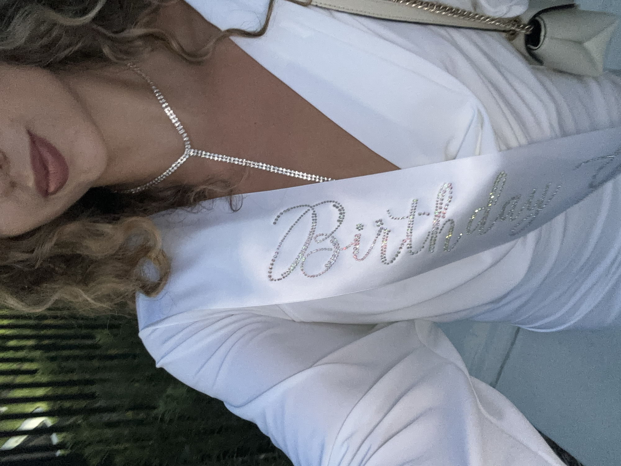 A medium shot captures a woman wearing a white dress and a white sash with "Bride" written in rhinestones. She has curly brown hair and is wearing a diamond necklace. The background appears to be outdoors with greenery and a fence, suggesting a location in Hallandale Beach, United States of America. A light-colored purse is visible on her right side.