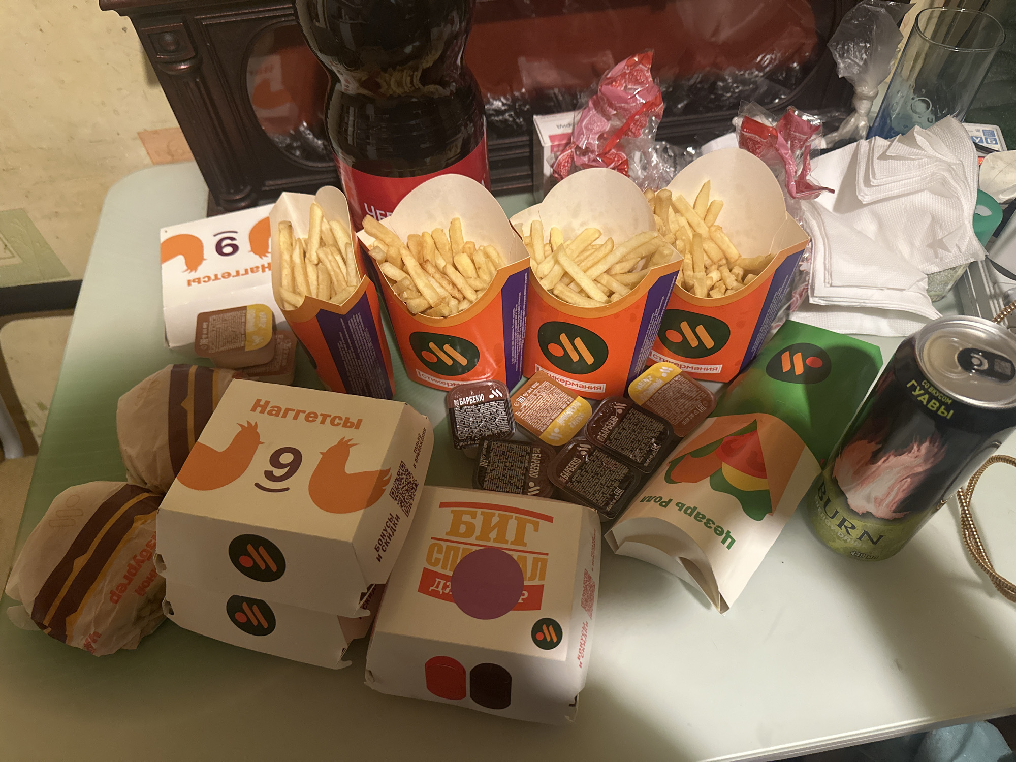 The image shows an assortment of fast food items, primarily from the Russian chain "Vkusno i tochka", a rebranded version of McDonald's. Visible are several containers of French fries, a box of 9 chicken nuggets, a "Big Special" burger, several smaller burgers, various dipping sauces labeled "BBQ", a "Caesar Roll" sandwich, a bottle of soda, a can of "Burn" energy drink with guava flavor, and napkins. Text on the packaging is in Russian. The location is likely Moscow, Russia, given the Russian language and the presence of a "Vkusno i tochka" restaurant meal. The scene suggests a meal either ready to be consumed or recently consumed, likely indoors on a table or other flat surface. No people are visible.