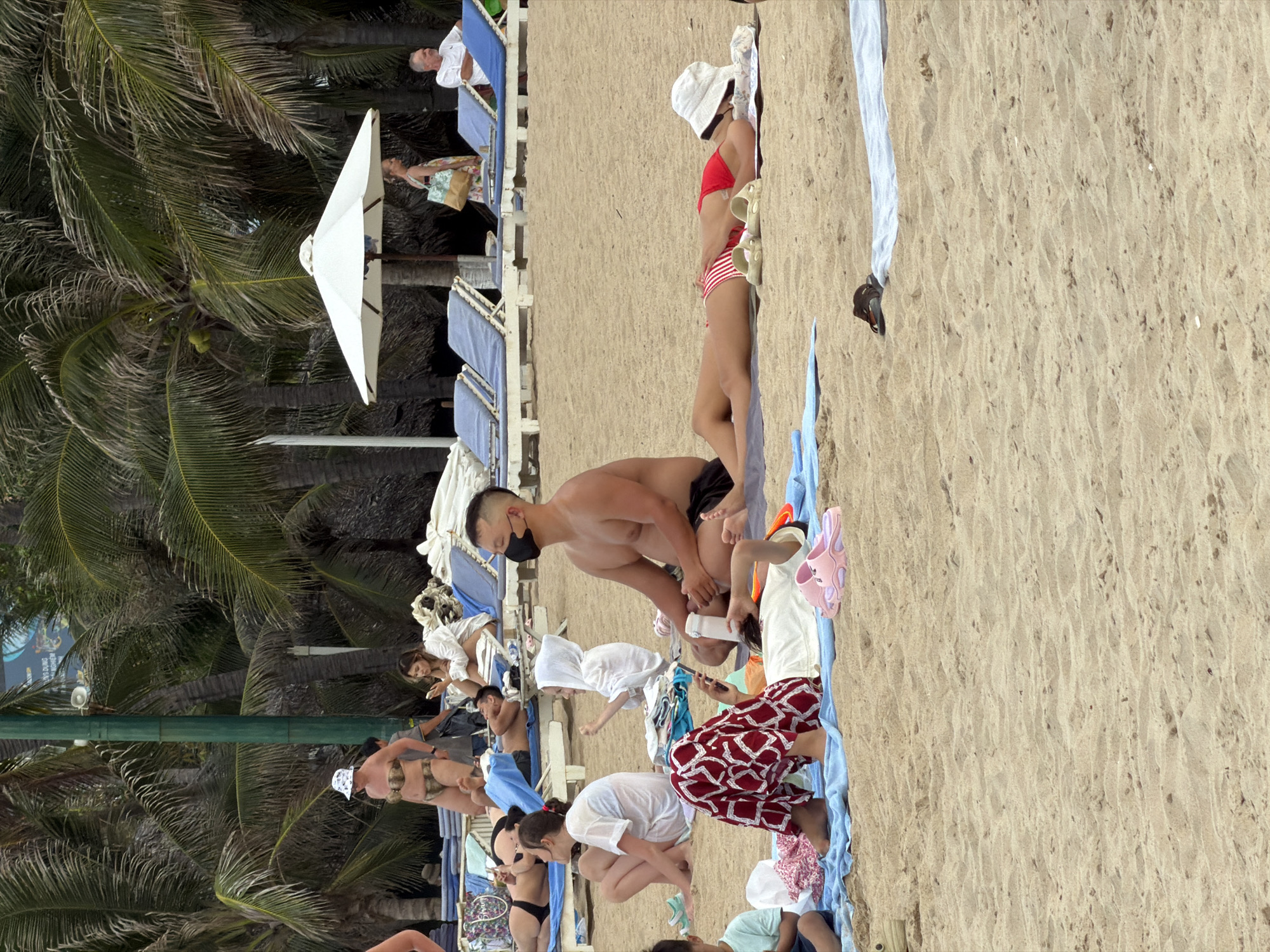 This image depicts a beach scene in Nha Trang Ward, Vietnam, on a sunny day. Numerous people are present, relaxing on lounge chairs and laying on towels on the sand. Palm trees line the background, and a white beach umbrella provides shade.
In the foreground, a person wearing red and white patterned pants is lying on a towel. To their right, another person in a red and white striped bikini is lying on a towel with a white bucket hat on their head. Next to them, a man wearing a black face mask is sitting upright on the sand, with his arms resting on his knees. A young child in a white hooded garment is sitting between him and the person in the patterned pants.
Further back, several people are visible on lounge chairs. Some are wearing swimwear, while others are dressed in casual beach attire. A woman in a white top and shorts is sitting on a lounge chair, looking at her phone. Another woman in a bikini is lying on a lounge chair. A man in a white shirt is sitting on a lounge chair in the background, and a woman with a patterned bag is walking behind a beach umbrella. The sand is golden and textured, with some small shells and debris scattered across it. The overall atmosphere suggests a leisurely day at the beach.