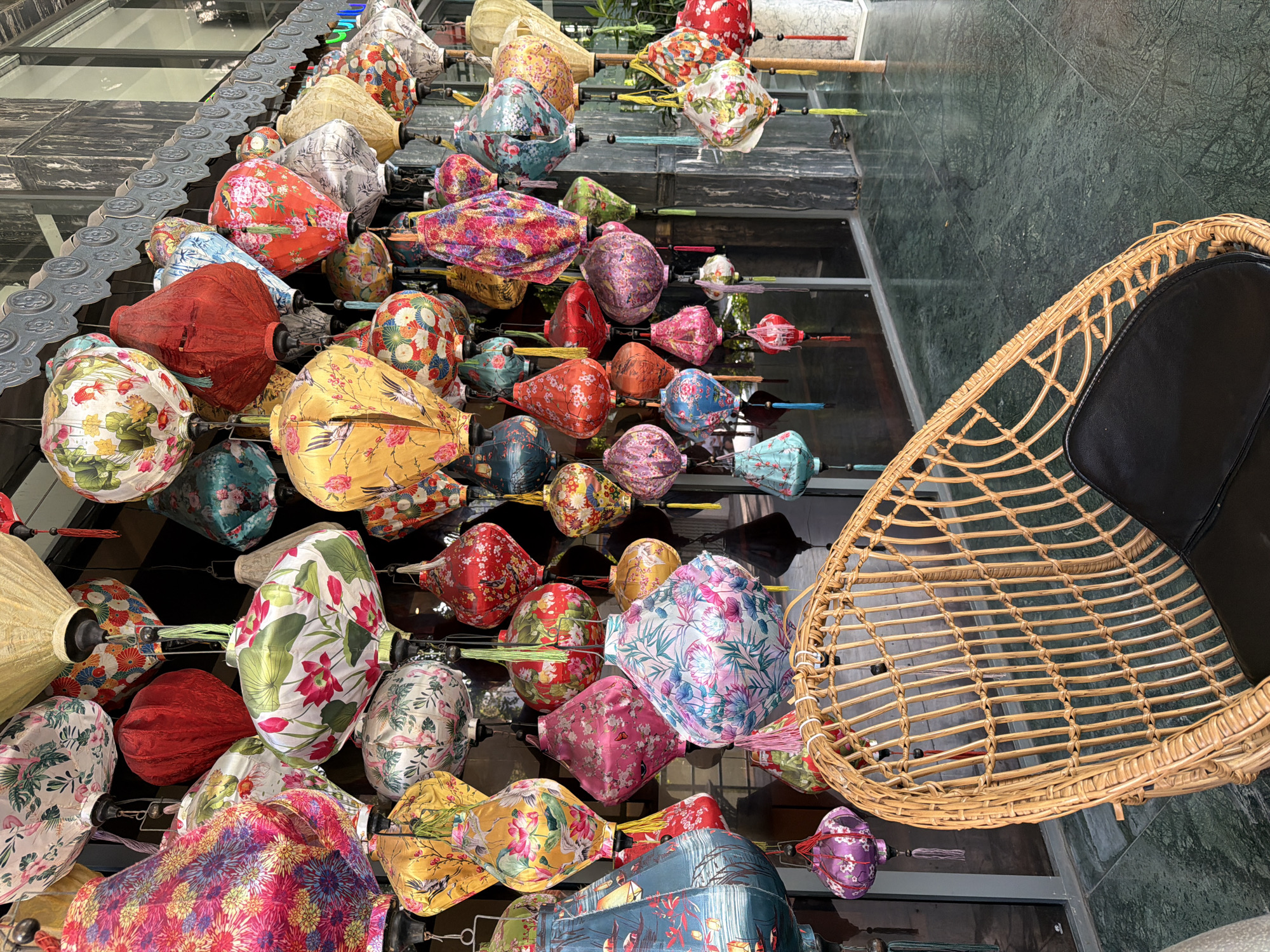 A large collection of colorful silk lanterns hangs from the facade of a building. The lanterns are of various shapes, sizes, and patterns, with floral designs and vibrant colors dominating. Some lanterns are lit, casting a warm glow. In the foreground, a wicker chair with a black cushion is placed on a green, marbled floor. The scene appears to be an outdoor seating area or a decorative display, possibly at a hotel, restaurant, or market. The lanterns are a common sight in Vietnam, often used for decoration during festivals and celebrations. The building's architecture and the style of the lanterns suggest a location in a Vietnamese city, such as Nha Trang, known for its coastal beauty and vibrant culture. The overall impression is one of festive ambiance and cultural richness.
