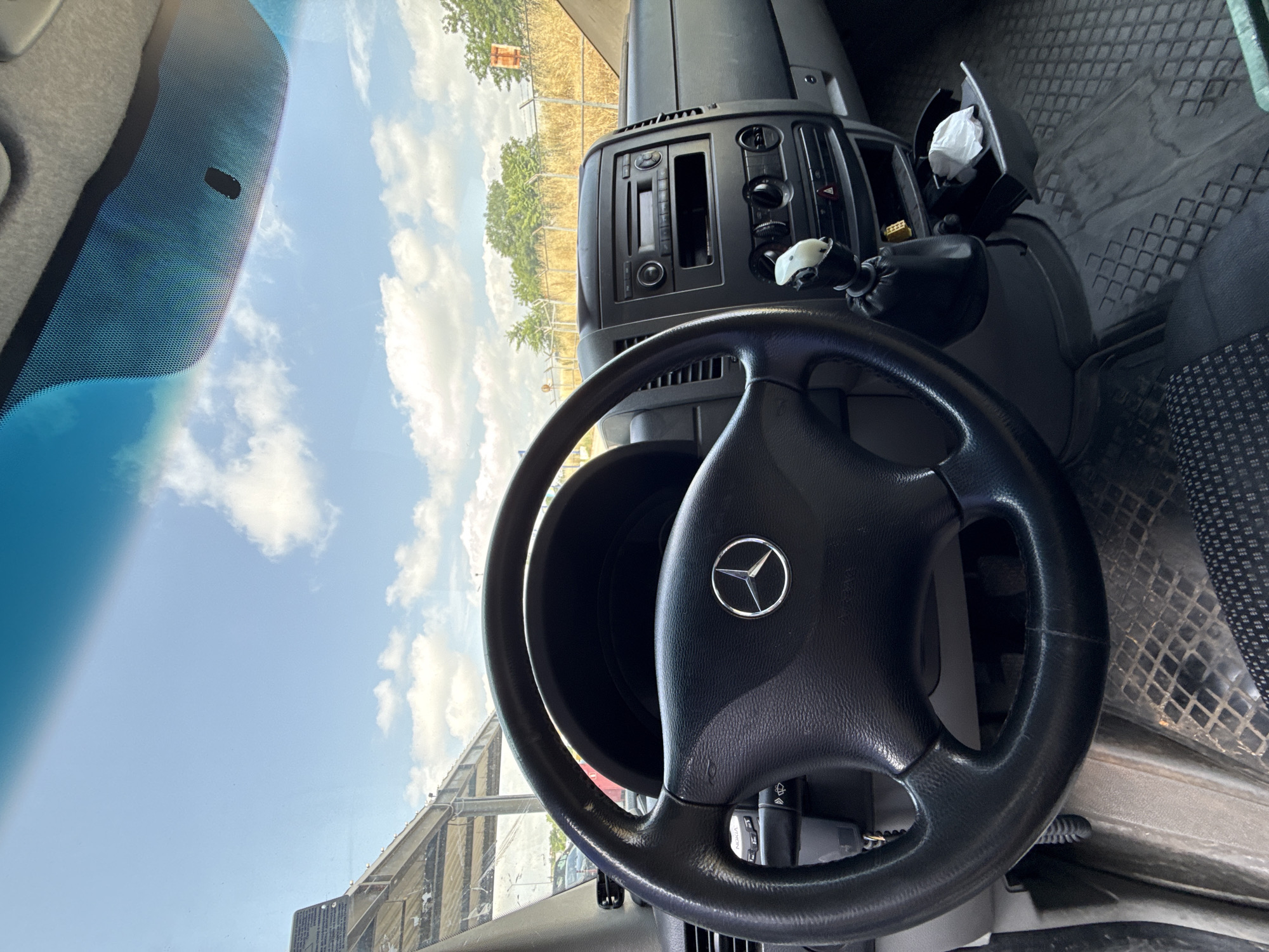 This media file captures the interior of a Mercedes-Benz vehicle, specifically focusing on the dashboard and steering wheel, viewed from the driver's perspective looking out the windshield. The image is rotated 90 degrees clockwise.
**Subjects:** The primary subjects are the vehicle's interior elements. A prominent black three-spoke steering wheel with a silver Mercedes-Benz logo occupies the center-right foreground. Behind it, the dashboard features a car radio/CD player unit, air conditioning vents, and various control knobs and buttons, including a red hazard light switch. To the right of the dashboard, a compartment holds a crumpled white tissue, and a black textured floor mat is visible on the passenger side. A phone holder or similar accessory, partially visible near the steering column, has the brand "Nokia" on it. Through the windshield, a vast blue sky with fluffy white clouds is visible, along with a landscape of dry, golden-brown vegetation and green trees in the distance. Part of an industrial or agricultural structure with metal framework is seen on the right, and a yellow marker or sign stands in the distant field.
**Activity or event taking place:** The vehicle appears to be stationary, likely parked outdoors, given the clear view of the sky and landscape. There is no visible human activity or event in progress.
**Notable details:** The scene is brightly lit, indicating it is daytime, likely during the summer or a warm season due to the dry vegetation. The sky is largely clear with scattered clouds. The vehicle is clearly identified as a Mercedes-Benz by the logo on the steering wheel. The interior suggests a functional, possibly commercial, vehicle like a van or a truck, consistent with the robust dashboard and textured floor. The overall impression is of a parked vehicle on a sunny day in an area with a mix of natural landscape and some infrastructure.
**Location cues:** The landscape with dry fields and the general appearance of the vehicle are consistent with a setting in Stara Zagora, Bulgaria.
**Visible text:** The word "Nokia" is visible on a device or accessory holder near the steering wheel.