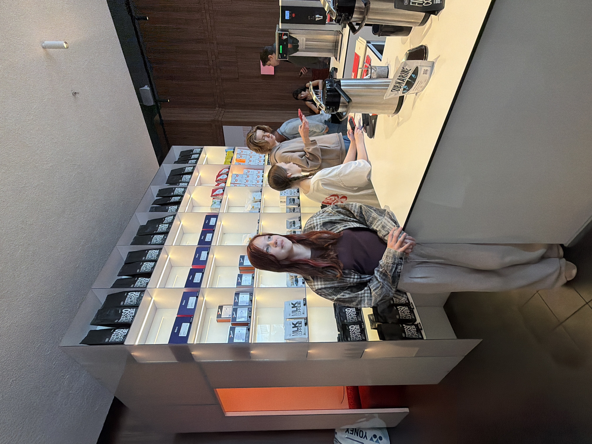 The image depicts the interior of a modern coffee shop, likely located in the city of Perm, Russia, given the context. In the foreground, a young woman with reddish-brown hair, wearing a plaid shirt over a dark top and light-colored trousers, stands facing right. She is positioned in front of a brightly lit retail display that showcases various packages of coffee, with "ESPRESSO COFFEE" visible on many of them. Further into the shop, behind a white counter, are several individuals. One young woman with blonde hair, wearing a light brown hoodie, is seated at the counter, possibly interacting with customers or staff. Another individual, a young man with blonde hair, stands behind her, looking at his phone. In the background, other patrons are visible, including a woman seated and looking at her phone, and a man standing and speaking with another person. The counter area is equipped with commercial coffee machines and a silver thermal carafe. The overall ambiance suggests a bustling but organized cafe environment.