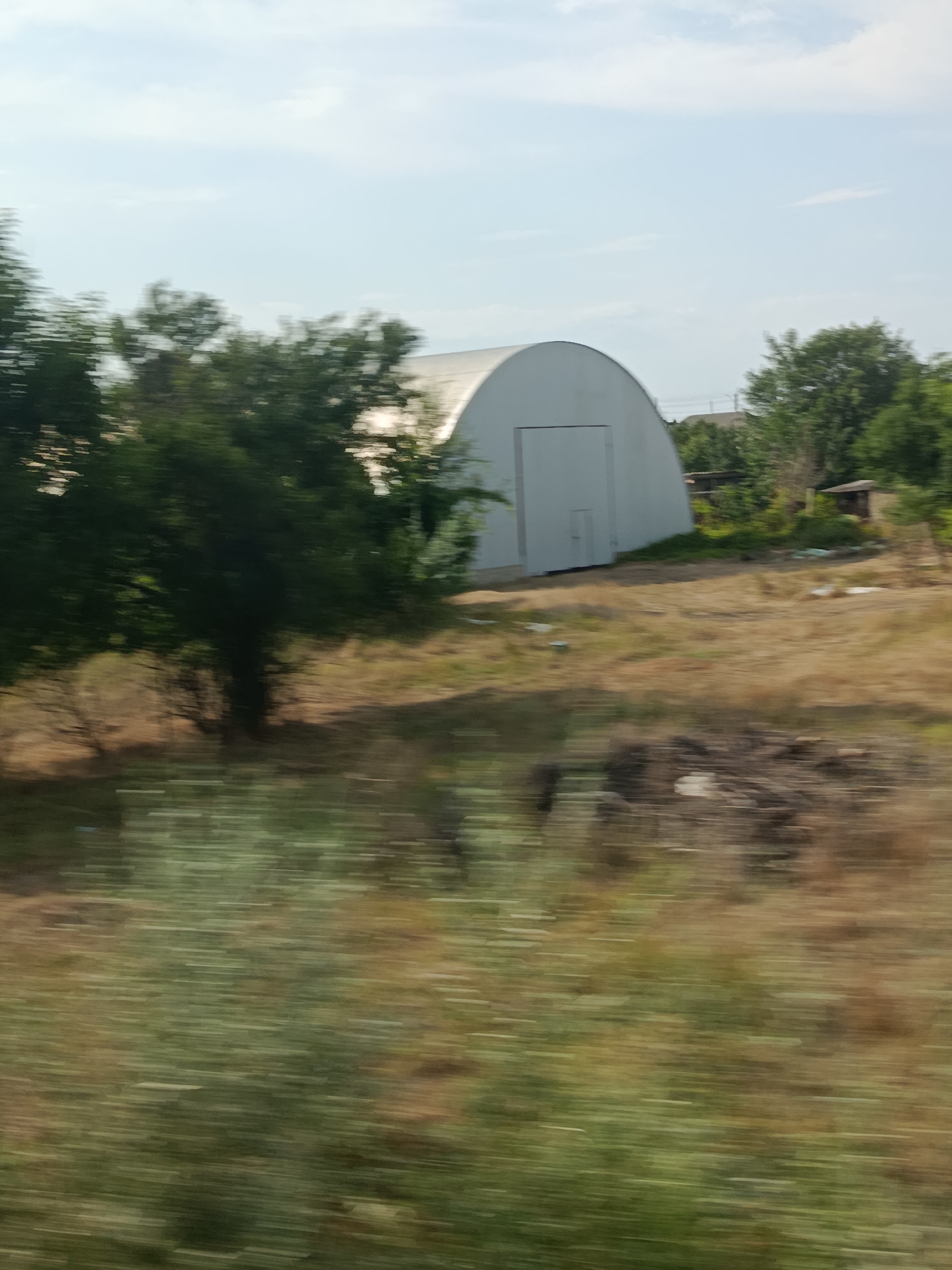 A motion-blurred image captures a large, white, Quonset hut-style building with a large, vertical door. It is situated in a dry, grassy field with scattered trees. The sky is light blue with some clouds, suggesting a daytime setting. The motion blur indicates that the photo was likely taken from a moving vehicle. The scene appears rural, with glimpses of other small buildings and trees in the background, hinting at a village or countryside setting. There is no visible text in the image.