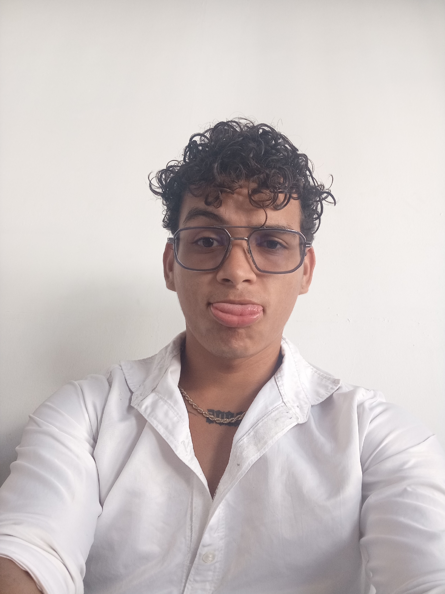 A close-up, waist-up portrait shows a young person with curly dark hair and glasses against a plain white wall. The individual is wearing a white button-down shirt and a gold chain necklace. Their tongue is sticking out to the side, and they are looking directly at the camera. The image appears to be taken in an indoor setting with neutral lighting. There are no discernible architectural features or identifying landmarks to confirm a specific location within Juan Griego, Venezuela.