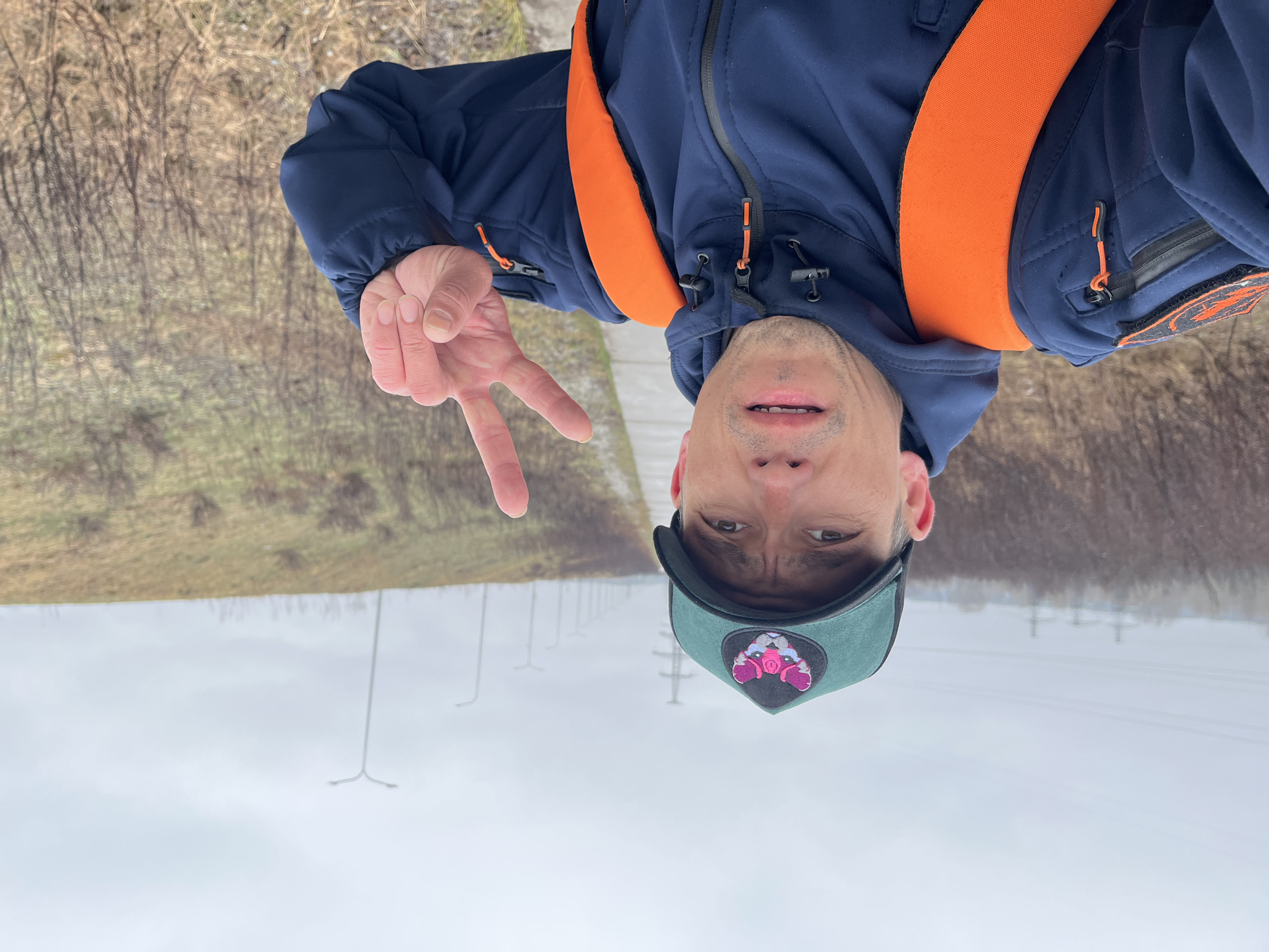 The image displays a man taking a selfie outdoors in what appears to be Chaikovsky, Russia, with the original image rotated 90 degrees. The man, seen from the chest up, wears a dark blue jacket accented with bright orange zippers and a prominent orange strap or harness over his shoulders. He has a dark green baseball cap featuring a distinctive pink and black logo, which resembles a stylized animal or character face. His left hand is raised, making a peace sign, and he has a neutral to slightly smiling expression on his face.
The background shows an overcast, light grey sky across the upper portion of the frame. Below this, on the right side, there is an area of dry, light brown grass and bare trees or shrubs, suggesting a late autumn or winter season. In the distance, several utility poles with power lines and streetlights are visible, lining what appears to be a road or pathway. The overall impression is a cool, cloudy daytime scene. There is no discernible text beyond the graphic logo on the cap.