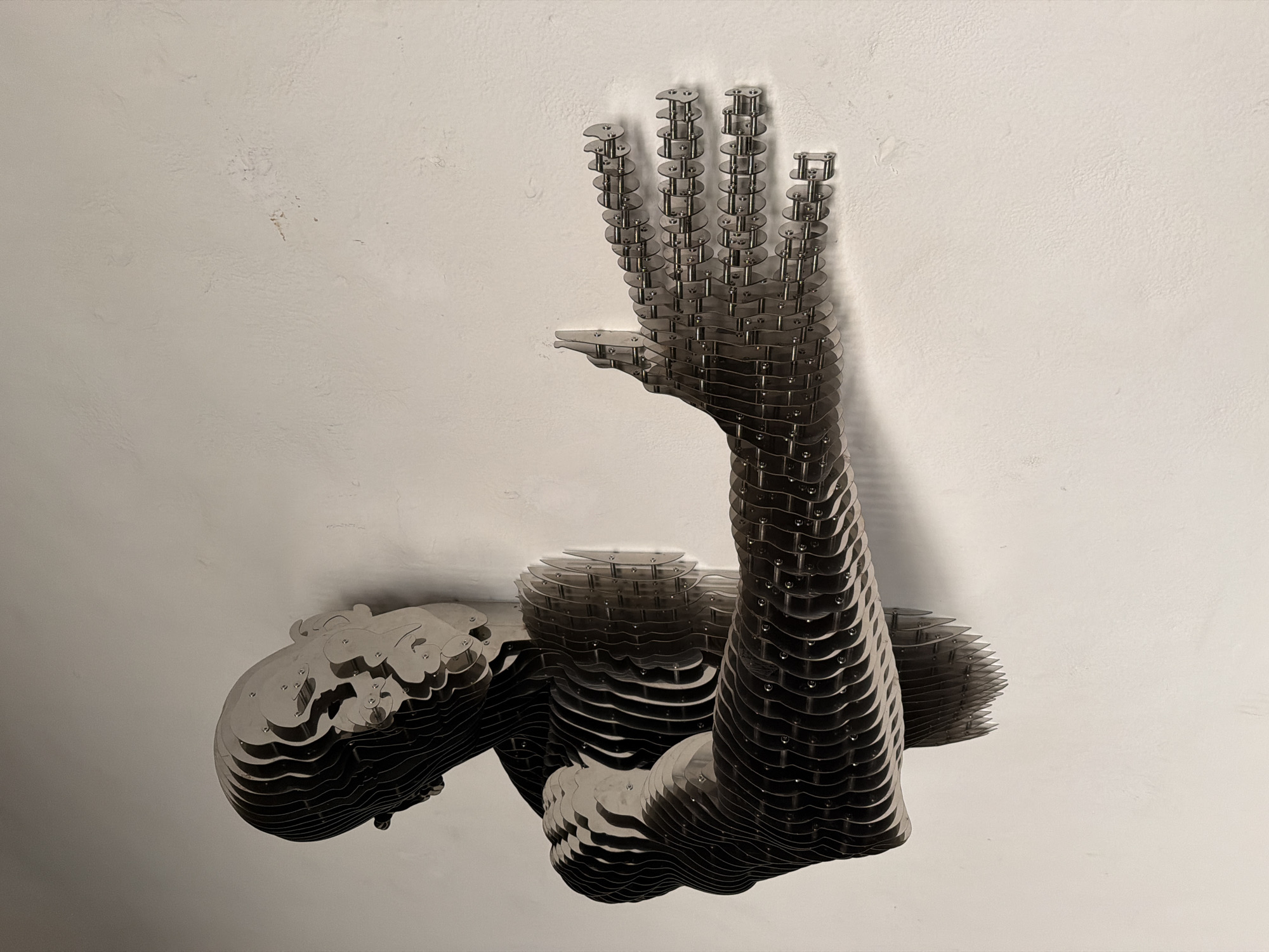 The image shows an abstract sculpture of a human figure attached to a light gray or white wall. The sculpture appears to be constructed from many thin, stacked metal sheets or plates, assembled to create the form of a reclining or contorted human body. The figure has a visible head, torso, arm, and hand. The hand is raised upwards in a gesture. The metal plates are held together with bolts or rivets. There is a shadow cast by the sculpture on the wall, indicating a light source from above or the side. The wall has a slightly textured surface. There are no other objects or people visible in the image. The image does not contain enough information to determine the location in San Bernardino Tlaxcalancingo, Mexico.