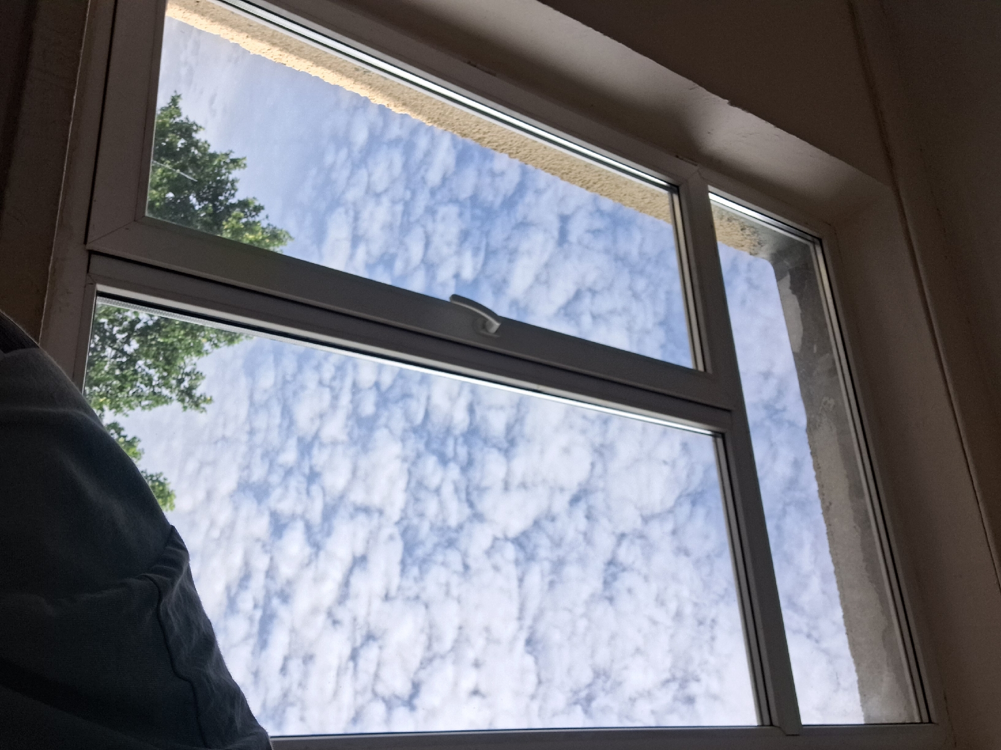 A view looking up and out through an open window. The window frame is white. The sky is filled with numerous small, puffy white clouds against a bright blue background. Green trees are visible below the window, partially obscuring the lower part of the sky. A person's shoulder and arm, wearing a dark grey garment, are visible in the foreground, partially obscuring the lower right side of the window. The wall around the window appears to be textured and a pale yellow color. The ceiling above the window is a neutral color and appears to have some rough texture. This scene is consistent with an apartment or house in Varna, Bulgaria, on a cloudy day.