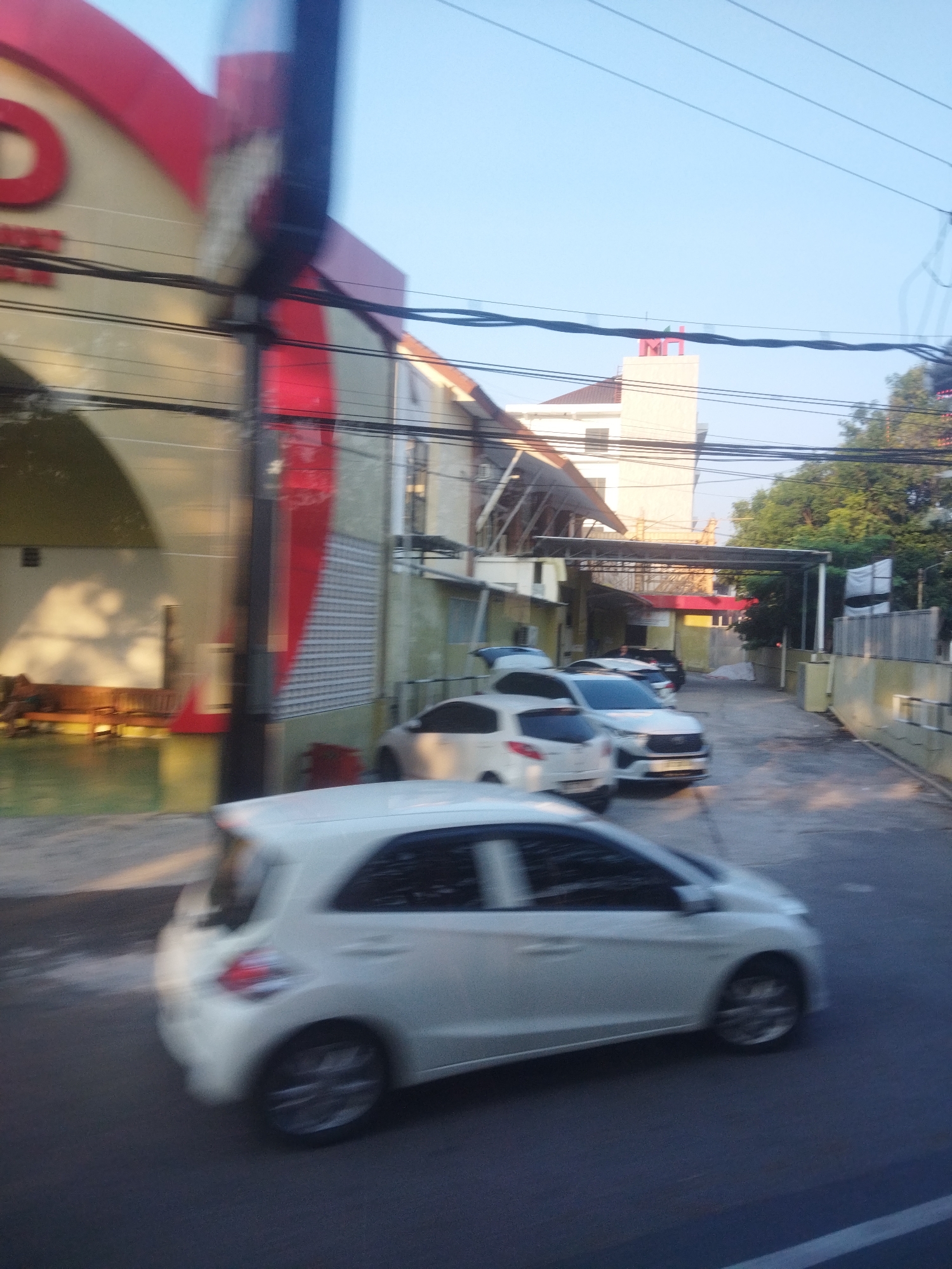 In Wonokarto, Indonesia, a blurry image captures a street scene with several white cars. In the foreground, a white hatchback is moving away from the camera, its rear visible. Further down the street, a white sedan is parked, and another white car is also visible. The background shows buildings with signs and awnings, and trees. Overhead, power lines are strung across the sky. The overall impression is of a typical urban street during daylight, although motion blur indicates the photograph was taken from a moving vehicle.