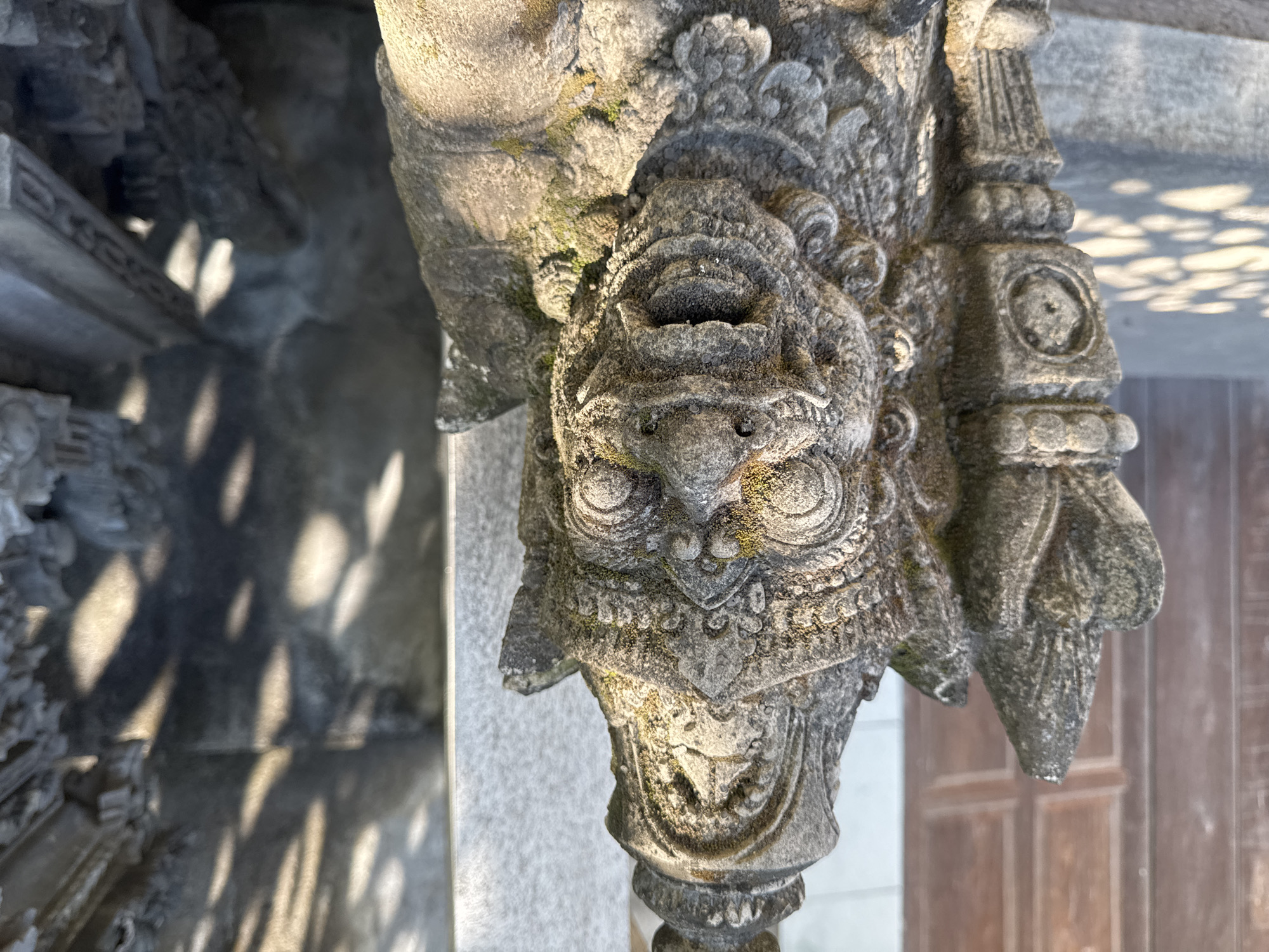 A close-up, eye-level shot shows a stone carving, likely a part of a building or temple, located in Kubu, Indonesia. The carving depicts a stylized, possibly mythical, creature with a prominent face and ornate details. The creature has large, circular eyes, a beak-like nose, and a patterned collar around its neck. The stone is weathered, with some moss or lichen growth. The background features architectural elements, including a wall with dappled sunlight filtering through an unseen structure, and a partial view of a wooden door or panel. The lighting suggests it is daytime.