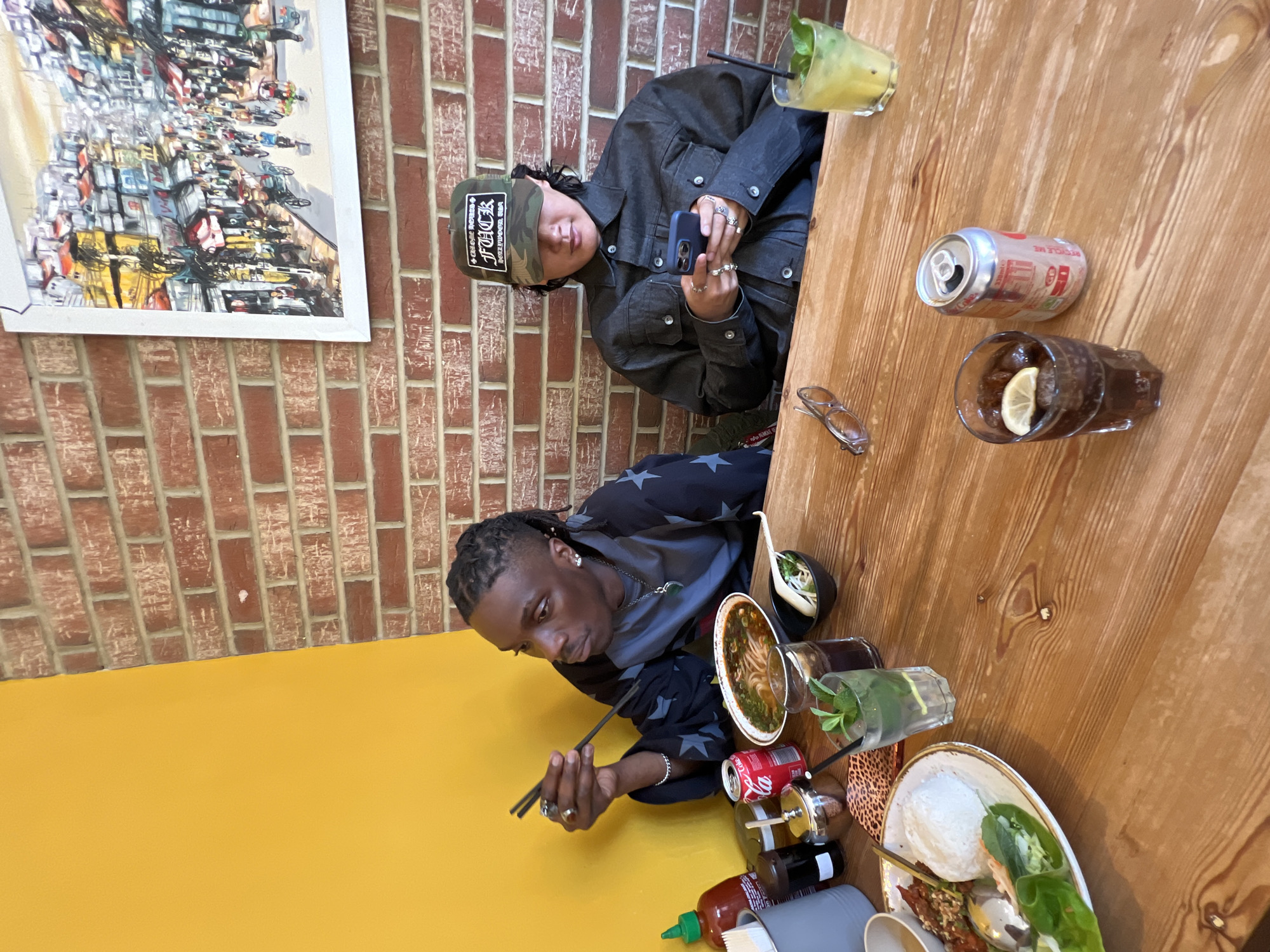 Two individuals are captured in a casual, indoor dining setting in London, United Kingdom. They are seated at a rustic wooden table, with one side of the room featuring an exposed brick wall and the other a bright yellow painted wall.
One person, positioned in the upper part of the frame, wears a dark grey denim jacket and a camouflage baseball cap with the prominent text "CRAZY POWER * F*CK * LOVE NO FEAR". They have multiple rings on their fingers and are engrossed in looking at a dark-colored smartphone. Adjacent to them on the table is a clear glass containing a light yellow, mint-garnished drink.
The second person, in the lower part of the frame, has dreadlocks and is wearing a dark top adorned with white stars. They are actively eating, holding chopsticks over a bowl of what appears to be pho or a similar noodle soup. On the table before them are various dishes, including a plate with white rice, fresh spring rolls, and grilled meat, along with a bottle of Sriracha sauce. Their drink, also mint-garnished, sits nearby.
Among the items on the table are two cans of Coca-Cola (one opened, bearing the text "RECYCLE ME"), a glass of iced tea with a lemon slice, and a pair of eyeglasses. A large, framed artwork depicting a dense, colorful cityscape hangs on the brick wall, adding a vibrant touch to the background. The overall atmosphere suggests a relaxed meal in a lively eatery, likely during daytime or evening given the good indoor lighting.