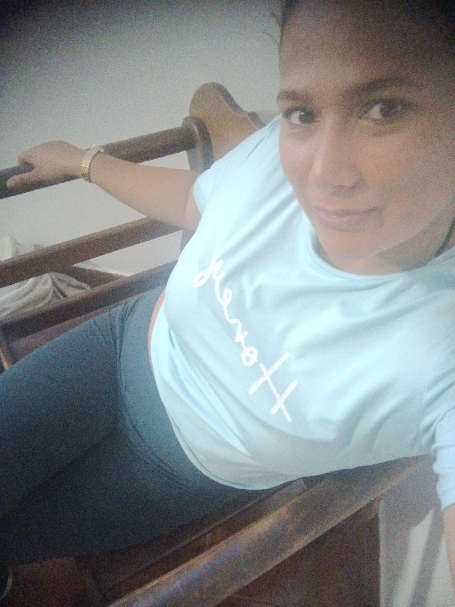 A woman is sitting on a wooden structure that appears to be a bunk bed or a loft bed, inside a room. She is wearing a light blue t-shirt with white text and dark-colored leggings. She has a wristwatch on her left wrist. The wall is plain and white. The picture is possibly taken indoors, with a soft light source. There is no evidence to suggest that the picture was taken in the city of Caracas, Venezuela.