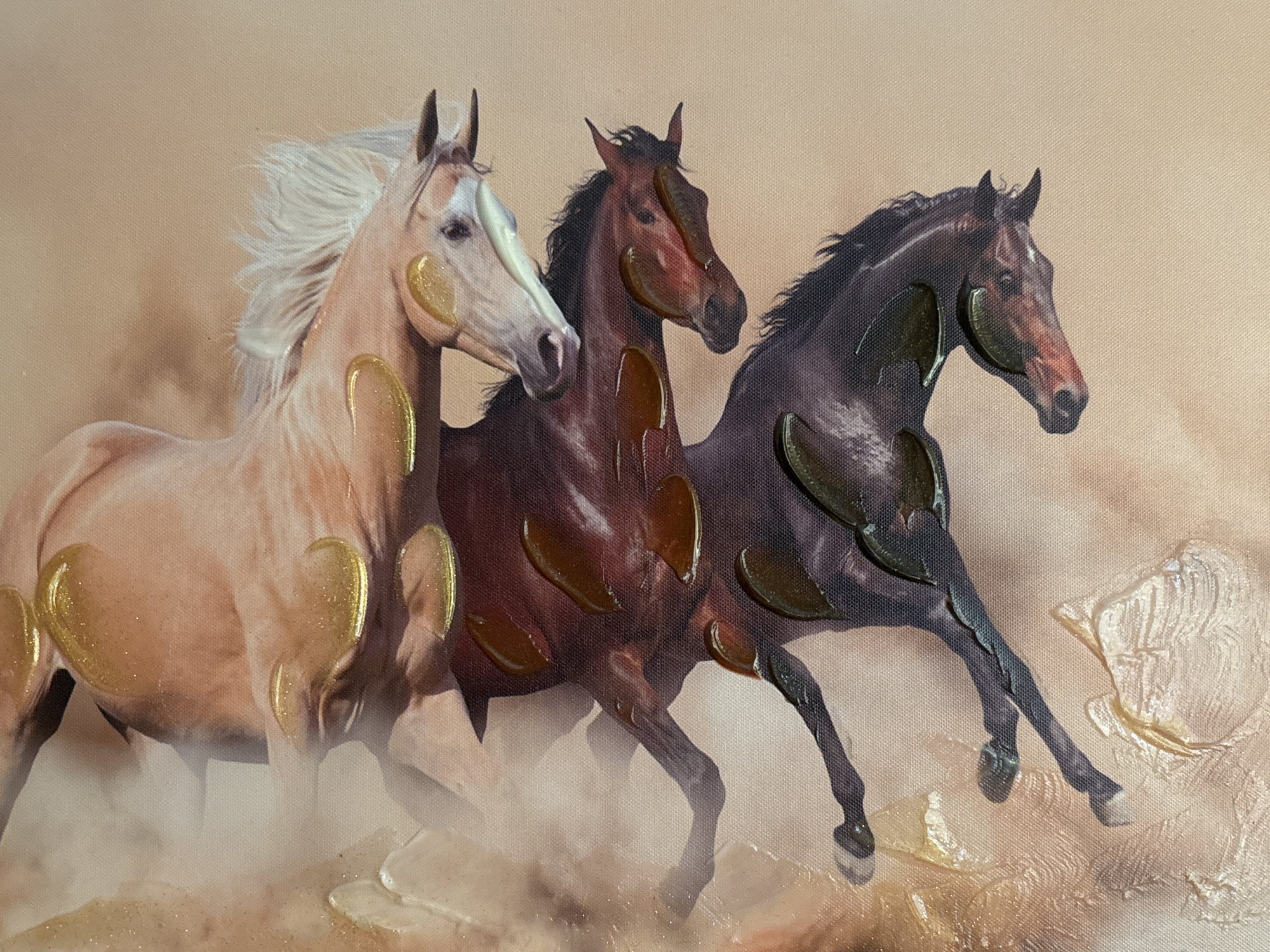 This image depicts a textured painting of three horses running. The horses are rendered in varying shades of brown and beige, with accents of gold and dark brown paint applied thickly, creating a dimensional effect. The background is a soft, peachy-brown color, suggesting a dusty or sandy environment. The horses are in motion, their manes and tails flowing, and dust appears to be kicked up around their hooves. The painting utilizes impasto techniques, with raised brushstrokes and dollops of paint, particularly noticeable on the horses' bodies and in the surrounding abstract elements. The style is expressive and somewhat impressionistic, focusing on capturing the energy and movement of the animals. The image is a close-up view of the artwork, with no discernible urban or natural landscape features visible, thus its location in Shumen, Bulgaria, cannot be determined from the visual information provided.