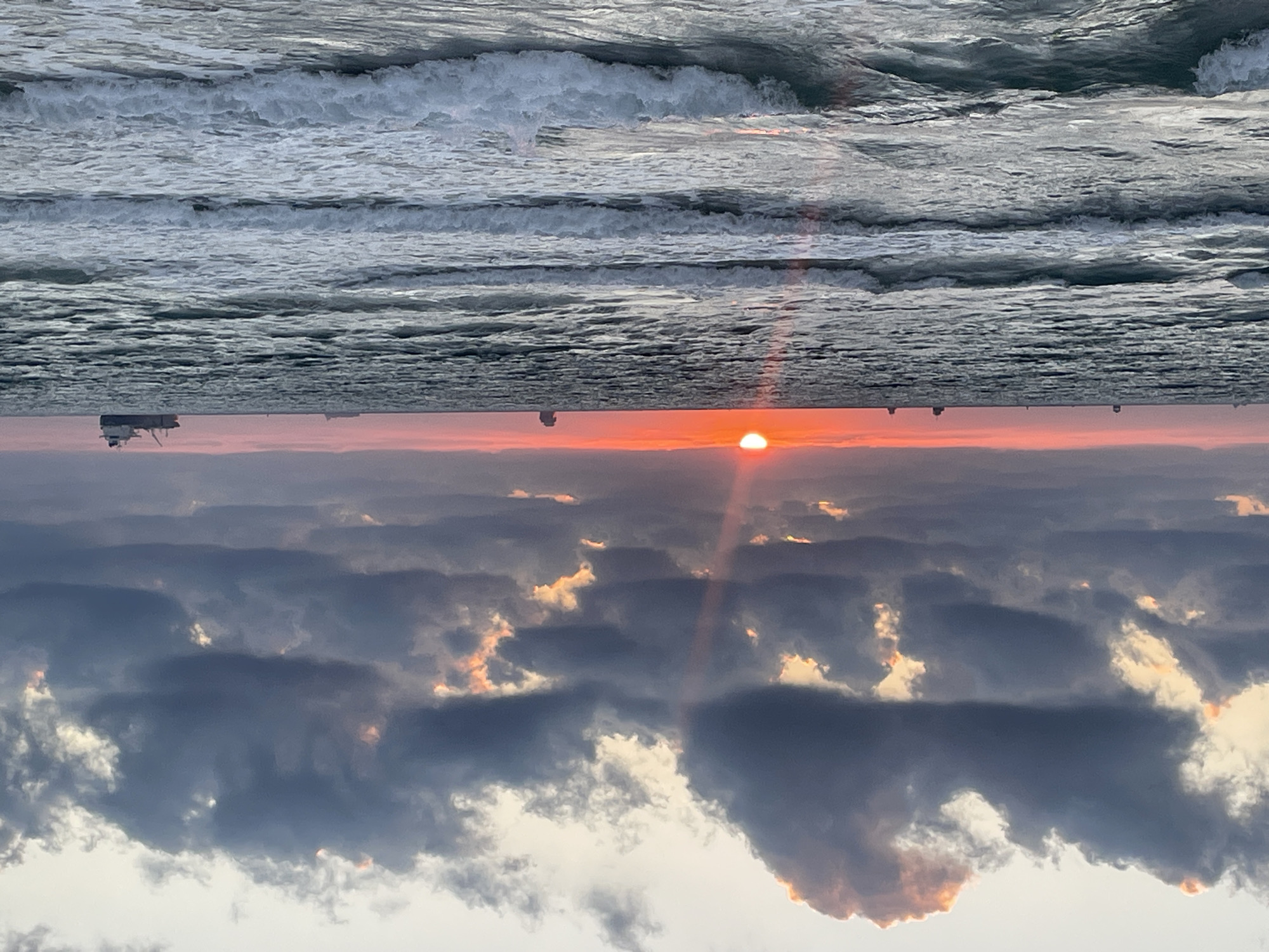 This is a photograph of a seascape during sunset. The image is oriented with the sky at the bottom and the sea at the top. The sea is a deep gray with white-capped waves. In the distance, on the horizon where the sea meets the sky, there are several small, dark shapes that appear to be structures or boats.
The sky is filled with dramatic clouds, colored in shades of pink, orange, and gray. The sun, a bright orange disc, is visible just above the horizon, casting a warm glow and a visible ray of light downwards. The clouds are illuminated by the setting sun, with some edges appearing bright orange and gold. The overall mood of the image is serene and dramatic, capturing the beauty of a coastal sunset.
The location is Haifa, Israel, and the time of day is sunset. There is no visible text in the image.