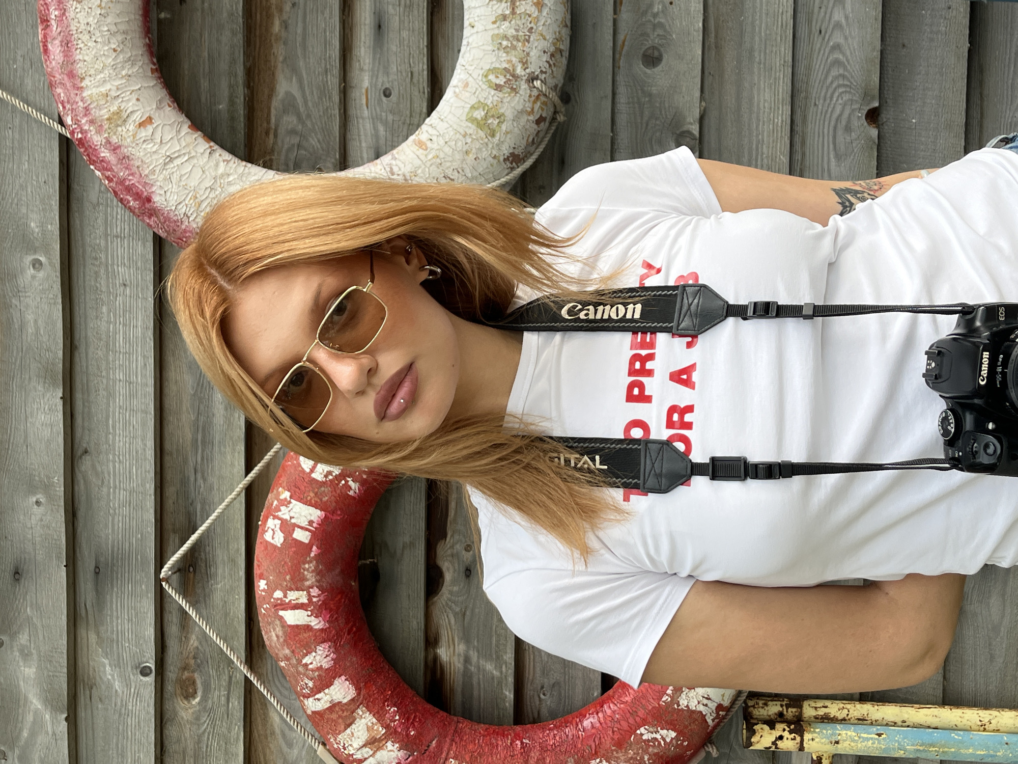 In this medium shot, a young woman with blonde hair and light-colored sunglasses rests against a weathered wooden wall. She is wearing a white t-shirt with red text that reads "TO PRETTY FOR A JUST..." and "DIGITAL". A black Canon camera strap with "Canon" printed on it is visible across her chest, attached to a black Canon camera partially visible on the right. Two life preservers, one white with some red and green markings, and another red with white markings, are positioned behind her and to her left, hanging from ropes. A thin rope is also visible, strung diagonally from the upper left to the lower center of the image. Her arms are crossed and resting on her chest. The lighting suggests it is daytime, likely outdoors, with the weathered wood and life preservers hinting at a seaside or poolside location, possibly a dock or pier. The overall mood is casual and relaxed.