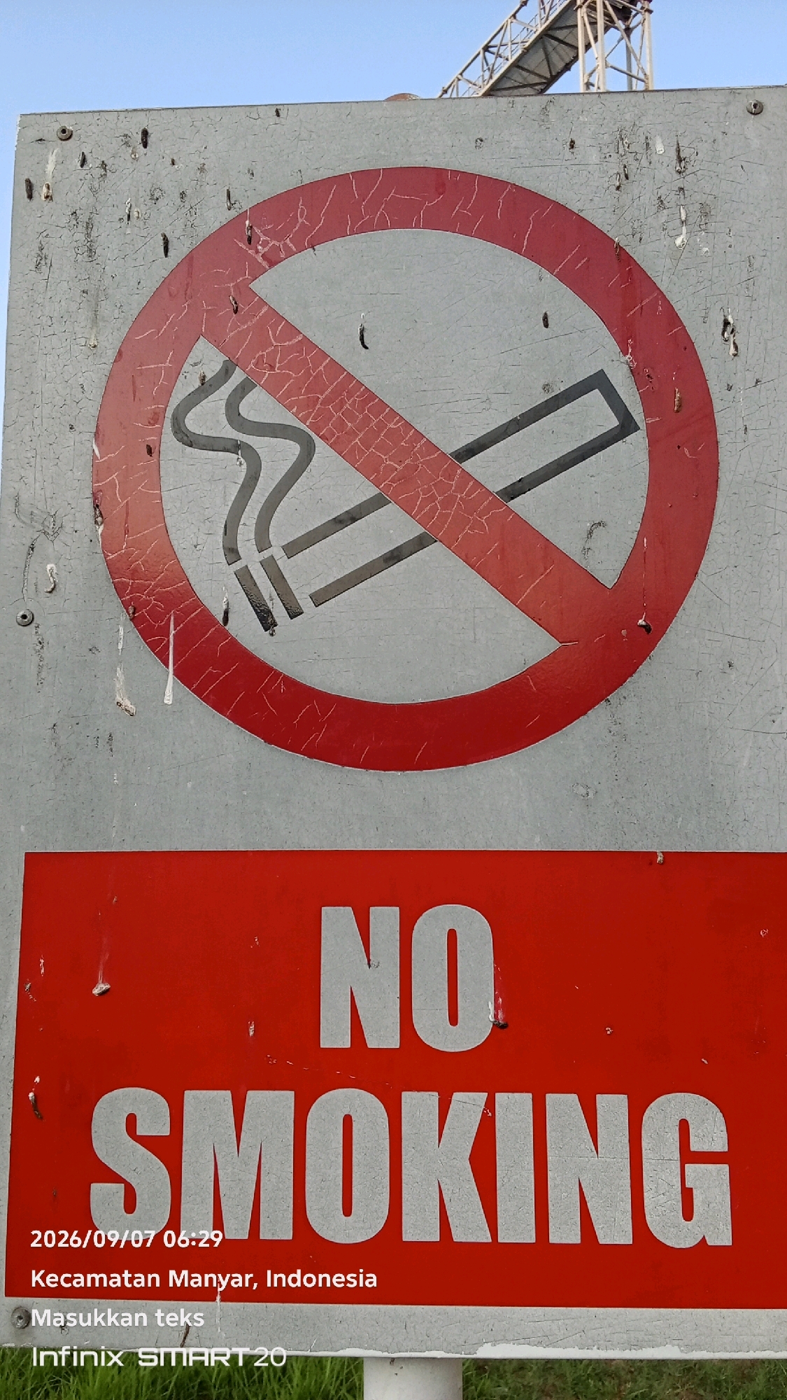 The image displays a "No Smoking" sign in a public outdoor area. The sign is predominantly gray with a circular red "no" symbol overlaying a depiction of a lit cigarette. Below the symbol, the words "NO SMOKING" are printed in large white letters on a red background. Additional text at the bottom of the sign indicates a date and time stamp (2026/09/07 06:29), the location "Kecamatan Manyar, Indonesia", and the text "Masukkan teks" and "Infinix SMART 20". The sign appears weathered, with peeling paint and some minor damage. The background shows a clear blue sky and hints of a metallic structure, possibly part of an industrial facility or bridge. The overall scene suggests an environment where smoking is prohibited, likely a public space such as a park, street, or entrance to a building. There are no people visible in the image.