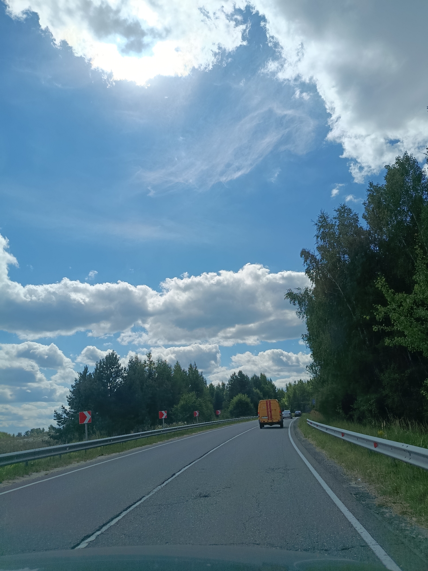 The image depicts a two-lane asphalt road in the countryside, likely on the outskirts of Gribchikha, Russia, under a bright, partly cloudy sky. The sun is visible through the clouds in the upper left portion of the frame. A metal guardrail runs along the right side of the road, and a similar guardrail is visible on the left side in the distance. Several trees, predominantly deciduous and coniferous, line both sides of the road. Red and white directional signs indicating a curve are posted along the left side of the road. A yellow van is traveling ahead on the right lane of the road, followed by at least two other vehicles. The road surface shows some signs of wear, with visible cracks and patches. The foreground suggests the image was taken from inside a vehicle.