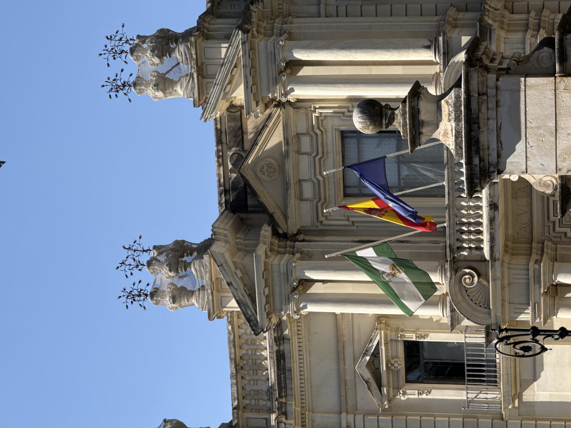 The image shows a close-up, low-angle view of the ornate facade of a historical building, likely in Seville, Spain, given the flags. The building is constructed of light-colored stone with intricate architectural details, including columns, carvings, and decorative moldings. Two flags are prominently displayed, hanging from flagpoles attached to the building. The upper flag appears to be the flag of Spain, with red and yellow stripes and a coat of arms. Below it is a flag with green and white stripes, which is the flag of Andalusia, a region in southern Spain.
The building's architecture suggests it could be a palace, a government building, or a religious institution. The style is Baroque or Neoclassical, characterized by its elaborate ornamentation and grandeur. The flags indicate a significant location, possibly a civic building or a place of cultural importance.
The time of day appears to be daytime, with bright sunlight casting distinct shadows on the facade, highlighting the three-dimensional relief of the carvings. The sky is a clear, vibrant blue, suggesting fair weather. There are also decorative elements resembling sculpted gargoyles or grotesques, covered in wire mesh, which might be a protective measure or an artistic addition. A wrought-iron lamppost with a decorative motif is also visible. Some stone elements of the building appear weathered, showing signs of age.
There is some faint text visible on the building, appearing as "FACV" on the right side, possibly part of a larger inscription. The overall impression is one of historical richness, architectural beauty, and regional pride, as evidenced by the display of the Spanish and Andalusian flags.
