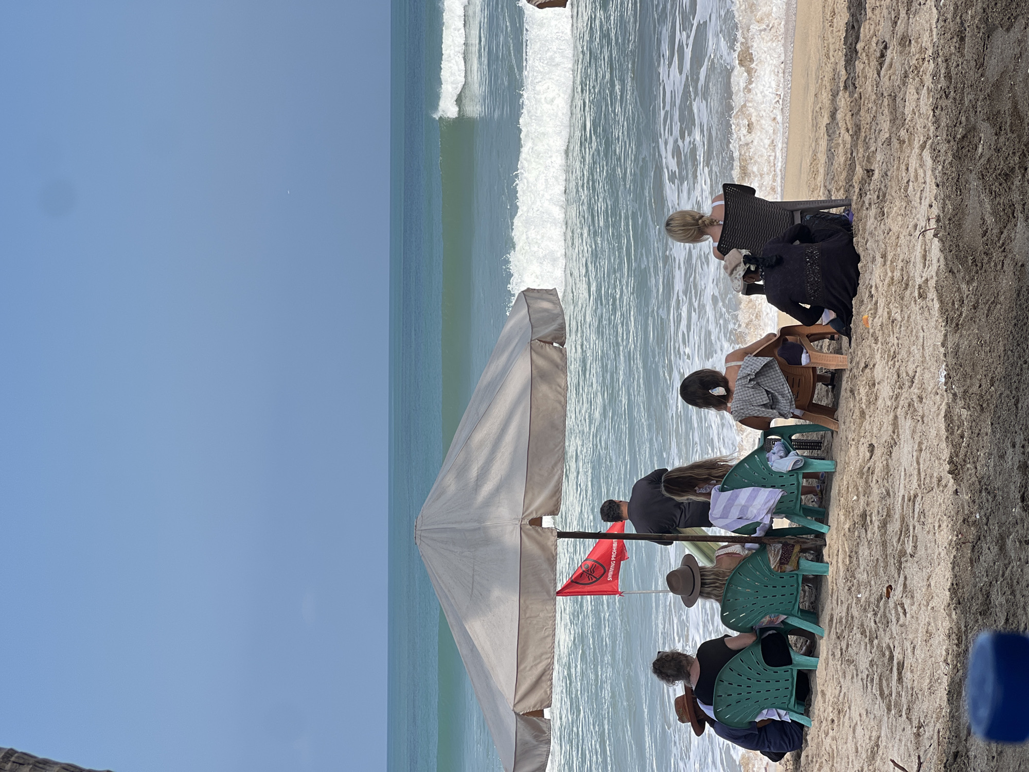 A group of people are sitting on chairs on a sandy beach facing the ocean. The waves are breaking in the distance. There is a large umbrella providing shade for some of the people. A red flag with a symbol is planted in the sand near the umbrella. The sky is clear and blue. The scene appears to be in Kuta, Indonesia.