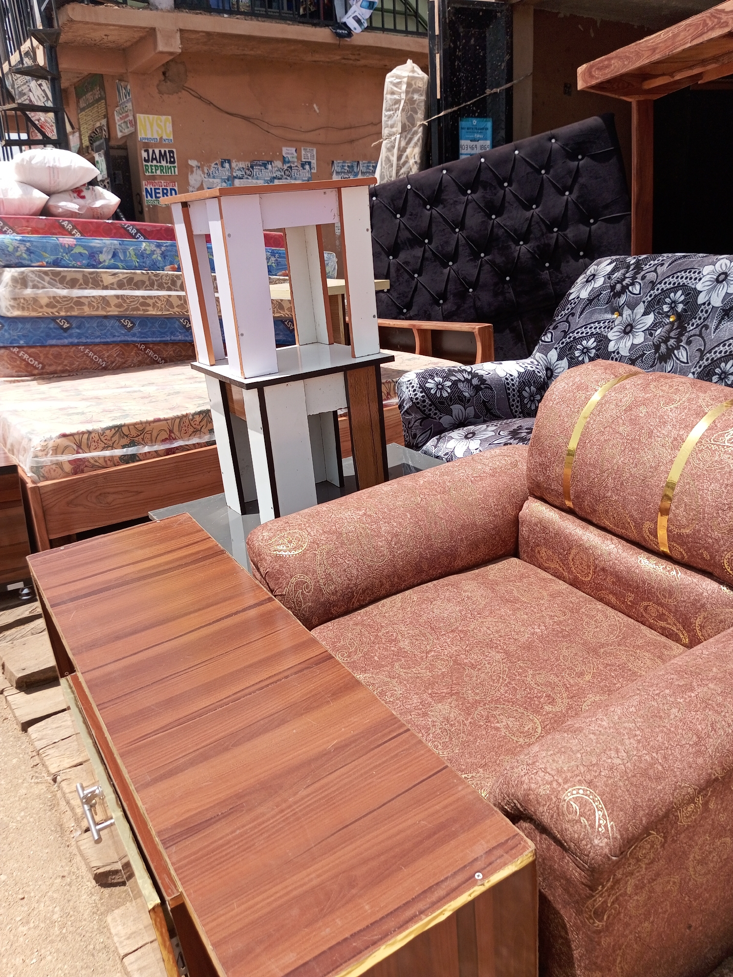 This media file shows an outdoor furniture and mattress shop in Bukuru, Nigeria. The scene is filled with various items, including stacked mattresses wrapped in plastic, a plush black tufted headboard, a patterned sofa, and a small white and wooden side table. In the foreground, a low wooden table with a wood-grain surface is positioned. The overall impression is one of a busy marketplace or retail space, with goods neatly displayed for sale.
The time of day appears to be daytime, with bright sunlight casting shadows. The weather seems clear. The setting is an urban environment, suggested by the building in the background with posters and signage, including a poster advertising "JAMB REPRINT" and "APPROVED CENTER NERD REGISTR". This indicates the location is likely a commercial area where students prepare for examinations. The presence of mattresses and furniture suggests a focus on household goods.
There are no people visible in the media file, but the arrangement of items implies commercial activity and sales. The overall mood is functional and business-oriented, with a focus on showcasing the products. The variety of items hints at a store catering to a range of customer needs for home furnishings and bedding. The textures and patterns of the fabrics on the furniture and mattresses add visual interest to the scene. The location cues, such as the signage and the outdoor display of goods, are characteristic of markets in many Nigerian cities.