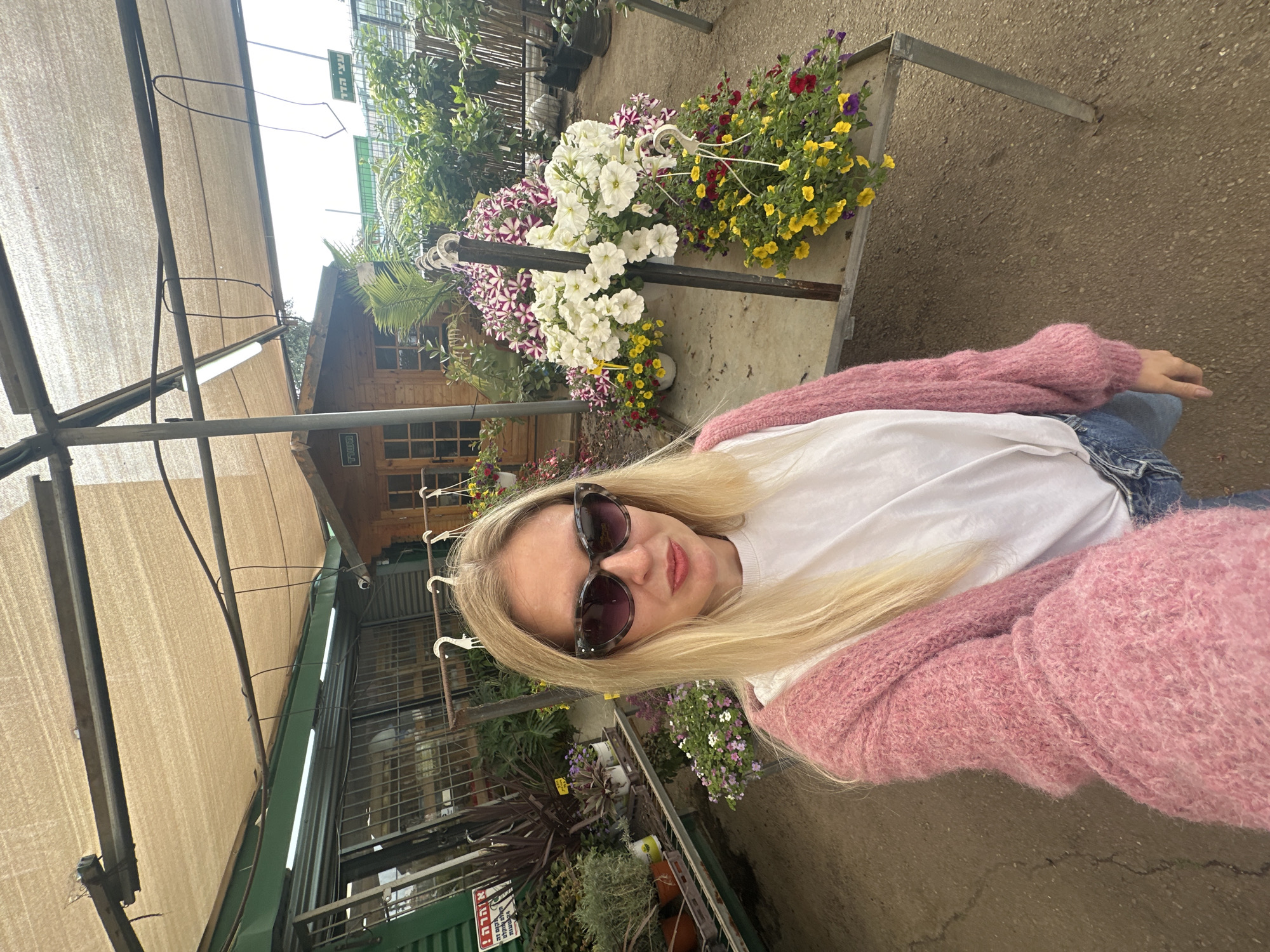 A woman in sunglasses and a pink sweater poses for a photo next to a flower stand. The woman is wearing a white shirt and jeans. The flower stand is filled with colorful flowers. In the background, there is a wooden building, a covered area, and various plants for sale. The picture was taken in a greenhouse or plant nursery, located in Petah Tikva, Israel.