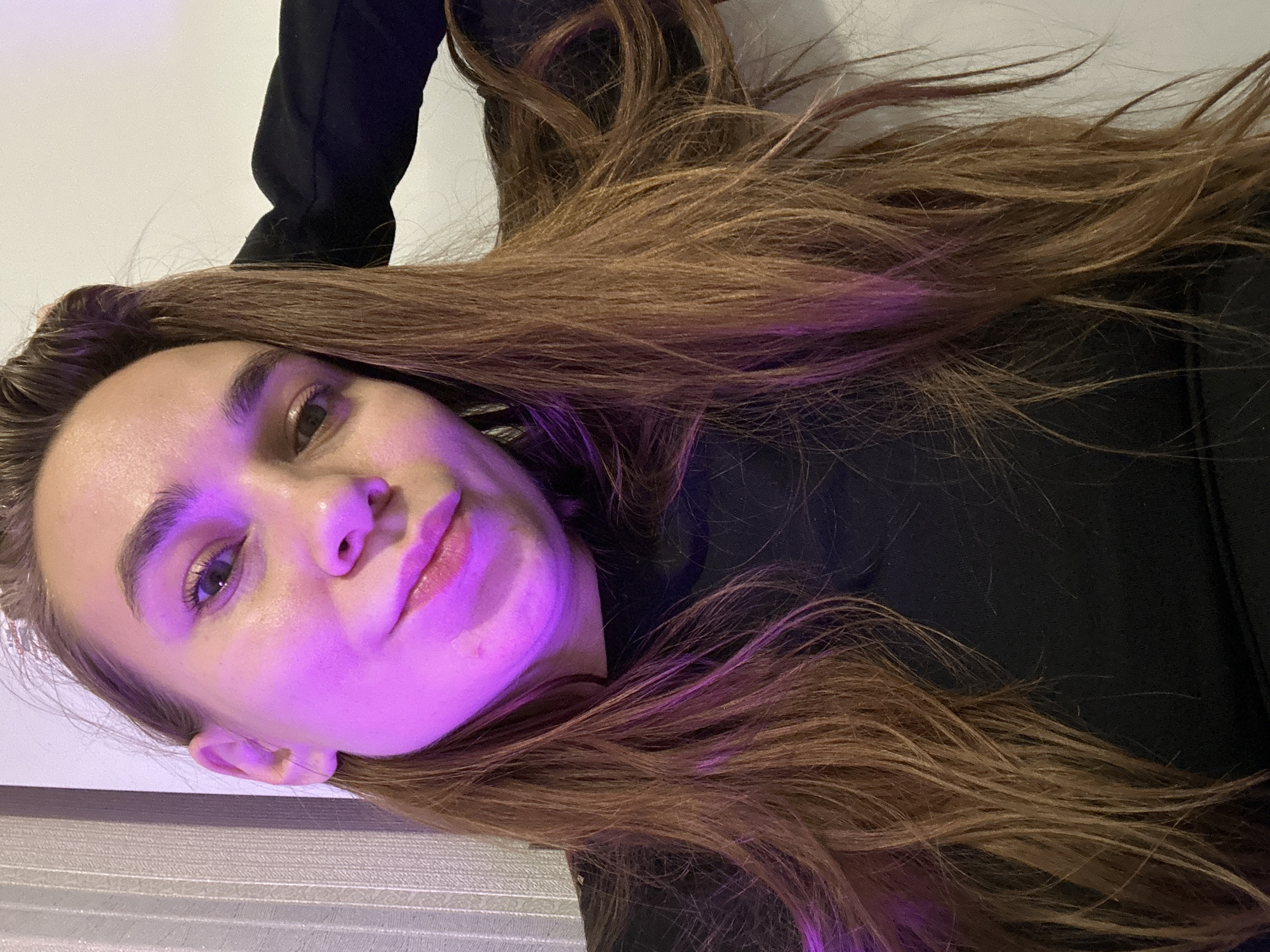A medium shot depicts a woman with light skin and brown hair. Her face is illuminated with a purplish-pink light. She wears a black shirt or jacket. The background is a neutral white. The location is not discernible from the image.