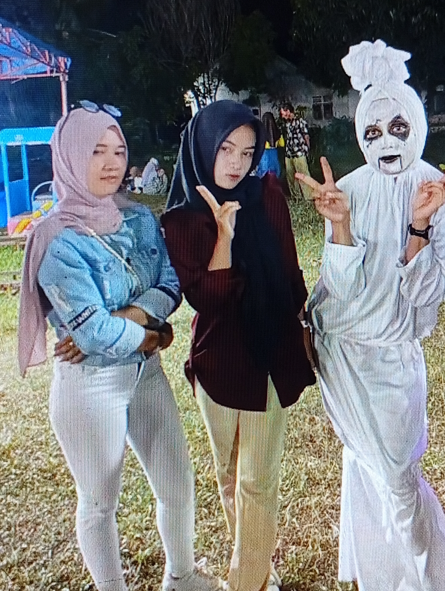 The image shows three individuals outdoors, likely in the city of Lhokseumawe, Indonesia, during what appears to be an event or gathering. In the foreground, from left to right, is a person wearing a light purple hijab, a light blue denim jacket over a white top, and white pants. They are wearing sunglasses on their head and have their arms crossed. Next to them is a person wearing a black hijab and a dark red shirt, with their arms in a peace sign gesture. To the right, a person is dressed in a white costume that resembles a ghost or specter, with face paint in black and white. They are also making a peace sign gesture. In the background, there are trees, some structures that might be part of a fair or event, and other people milling around. The ground is covered in dry grass. The lighting suggests it is evening or nighttime.