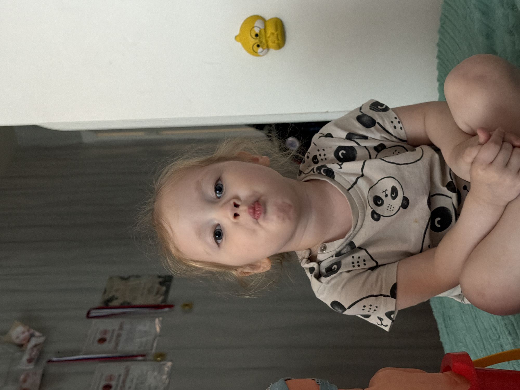 A young child, approximately two to three years old, with light-colored hair and blue eyes, is seated centrally on a teal-colored textured fabric surface, possibly a blanket or rug. The child is looking directly at the camera with pursed lips. They are wearing a light-colored short-sleeved t-shirt patterned with black panda bear designs. Their hands are clasped together in their lap, and legs are crossed.
To the child's left, in the background, a light-colored wall with vertical paneling is visible. Several framed documents, possibly certificates or awards, with red and white ribbons or medals, are hanging on this wall. A small figurine, appearing to be a doll, is also visible near the top left. Partially visible in the far left foreground is a red and orange plastic object, likely part of a toy.
To the child's right, a large, plain white vertical surface, resembling a wall or a large appliance, occupies the background. A yellow, anthropomorphic character figurine with large eyes and a downturned mouth, resembling a duck or bird, is affixed to this white surface. The scene is indoors, in what appears to be a residential setting in the city of Perm, Russia.