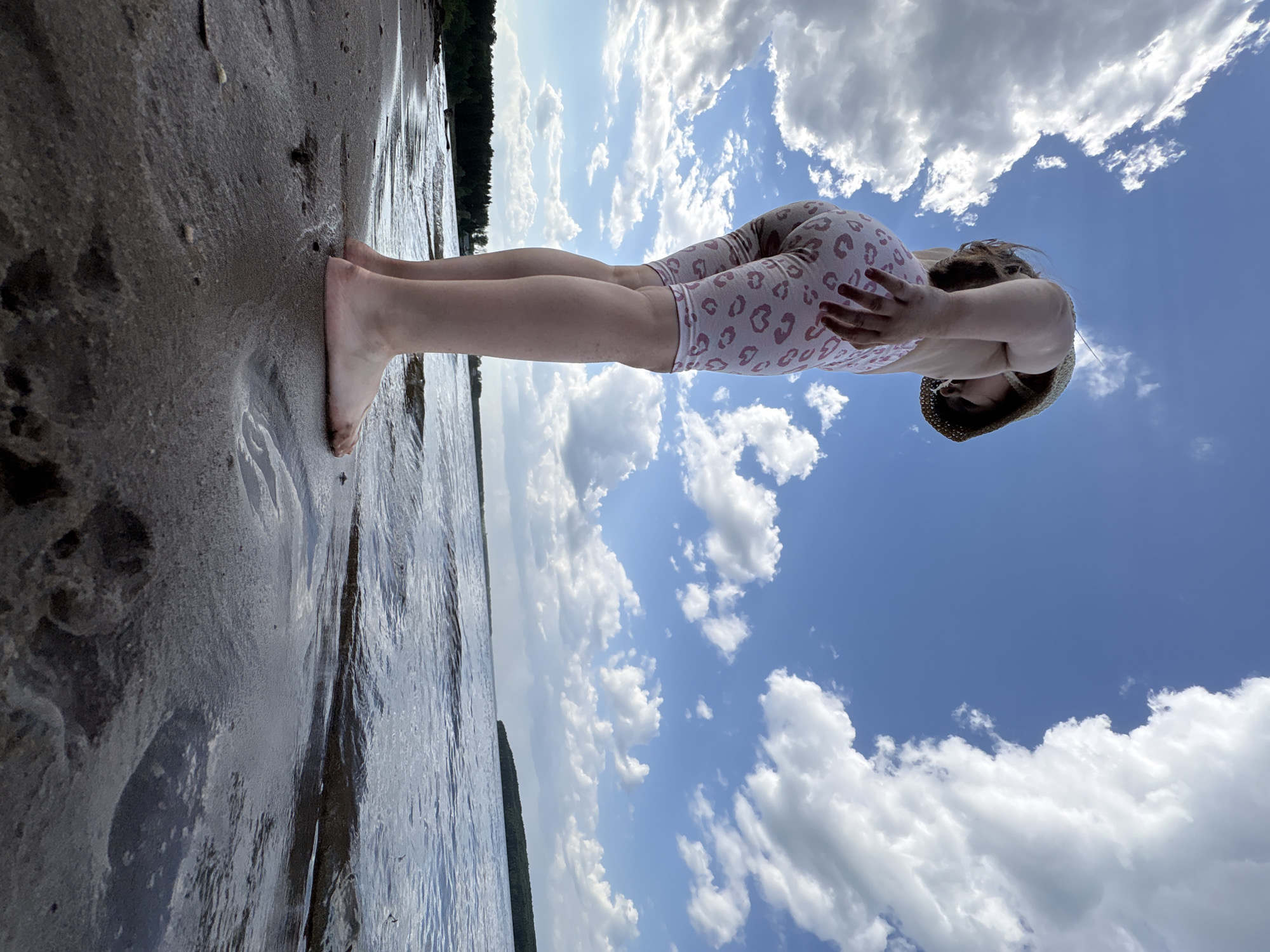 A young person in a light-colored leopard-print outfit and straw hat stands on a dark, wet beach. The person's feet are in the water. The sky is blue with puffy white clouds. Trees are visible in the background. The image appears to be taken at a low angle and rotated. The location is possibly Chaikovsky, Russia, based on the prompt.