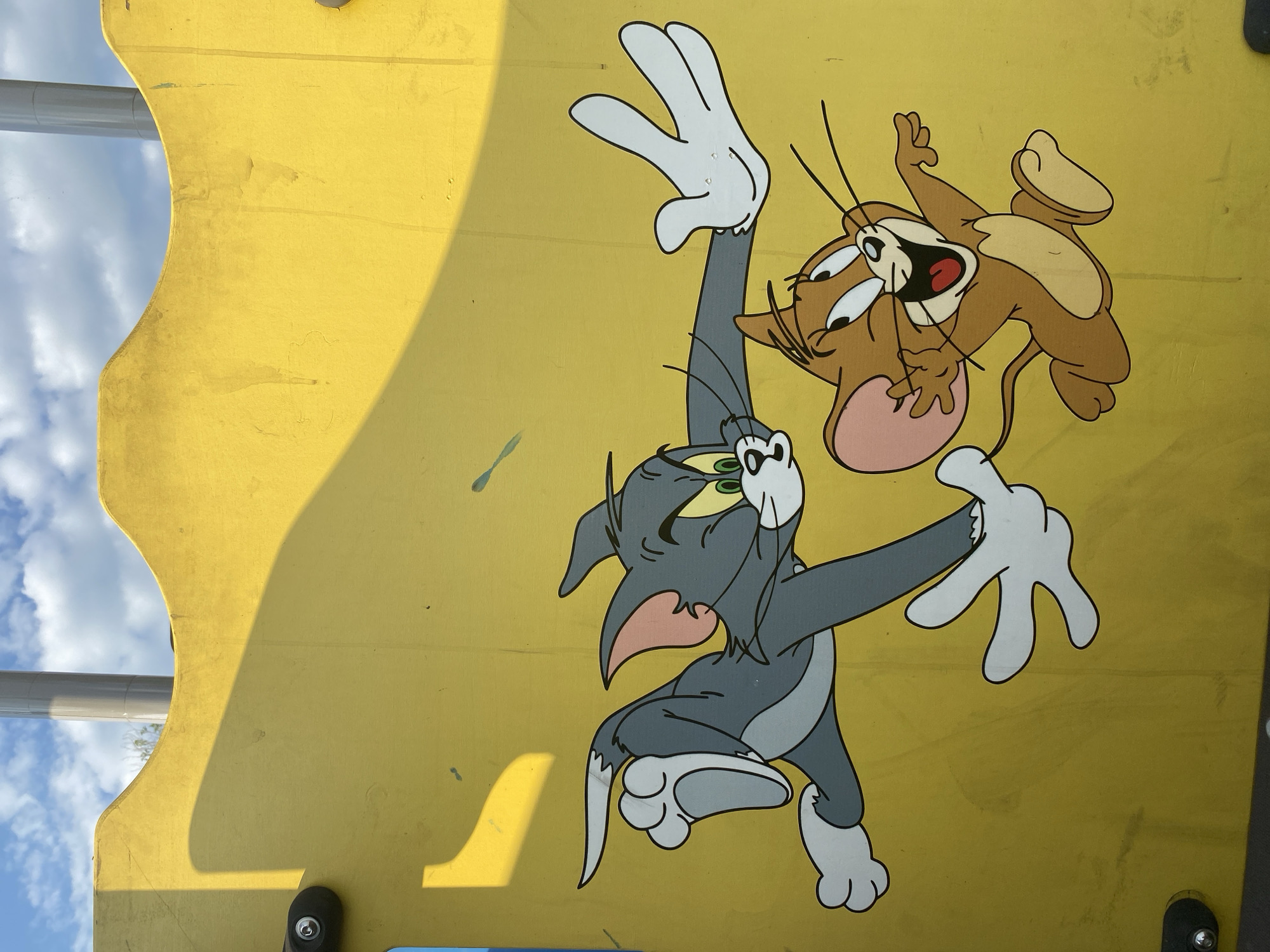 The image depicts a section of a yellow, outdoor playground structure with a cartoon mural of Tom and Jerry. Tom, the grey cat, is shown in a running pose with his body extended and his arms outstretched. Jerry, the brown mouse, is leaping dynamically with his mouth open in a joyful expression. The artwork is printed or painted onto the yellow surface, which shows some signs of wear and tear, including scuff marks and faint streaks. The top of the structure has a curved edge, and a shadow is cast across the surface from an unseen object, possibly a tree or another part of the playground equipment. In the background, a portion of a cloudy blue sky is visible, along with a light-colored pole, likely part of the playground's infrastructure. The location is identified as Petropavl City Administration, Kazakhstan.