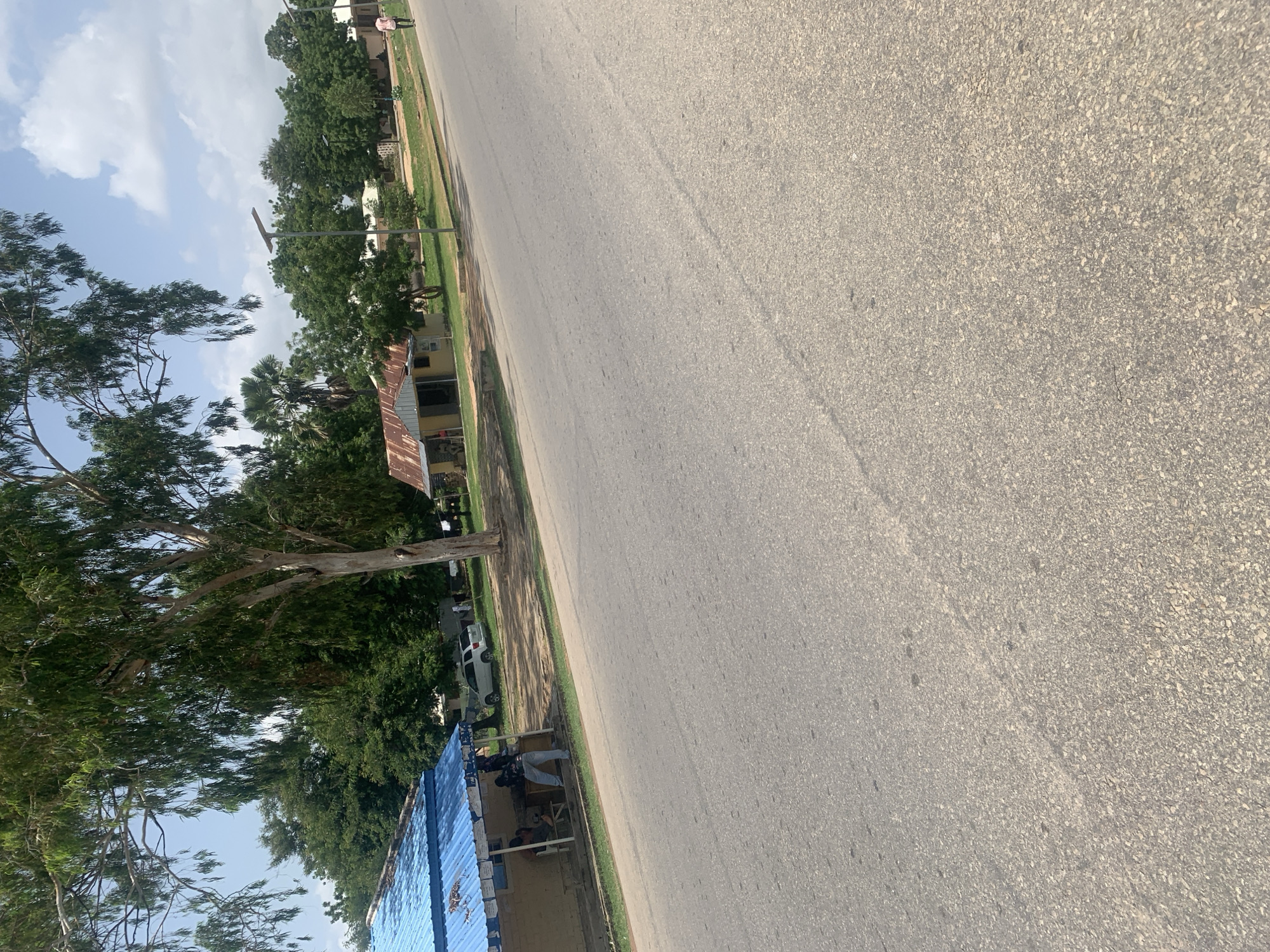 The image shows a tilted view of a street scene in Jalingo, Nigeria. A paved road takes up a significant portion of the frame, with a grassy verge and trees bordering it. On the verge, several small buildings with corrugated iron roofs are visible. One building has a blue corrugated metal roof.
A few people are present, some sitting under the blue-roofed structure, possibly engaged in a transaction or socializing. A white vehicle is parked on the side of the road, near the buildings. A tall tree with sparse foliage stands prominently on the left side of the image, its branches extending towards the sky. Beyond the buildings and trees, more greenery and structures can be seen in the background, suggesting a semi-urban or suburban environment.
The sky is partly cloudy, indicating a bright day, likely during daylight hours. The overall impression is one of everyday life unfolding on a street in a Nigerian town. No text is visible in the image.