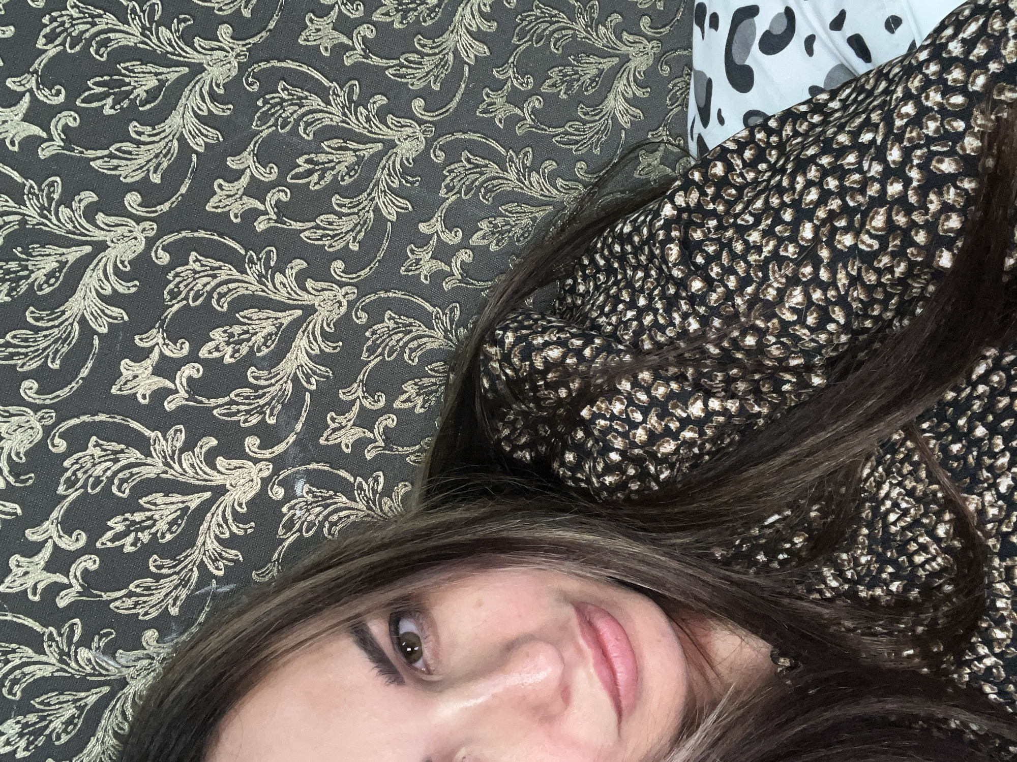 The image shows a close-up, selfie-style shot of a woman's face and upper body. She has long, dark brown hair with lighter streaks, brown eyes, and light-colored skin. She is wearing a black top with a small, dark brown and white animal print pattern. The background is a dark grey wallpaper with an ornate, scrolling floral pattern in a lighter, metallic gold color. To the right, a portion of a white pillow with a black and gray spotted pattern is visible. The lighting suggests an indoor setting. The woman is positioned on the left side of the frame, with her head tilted slightly.