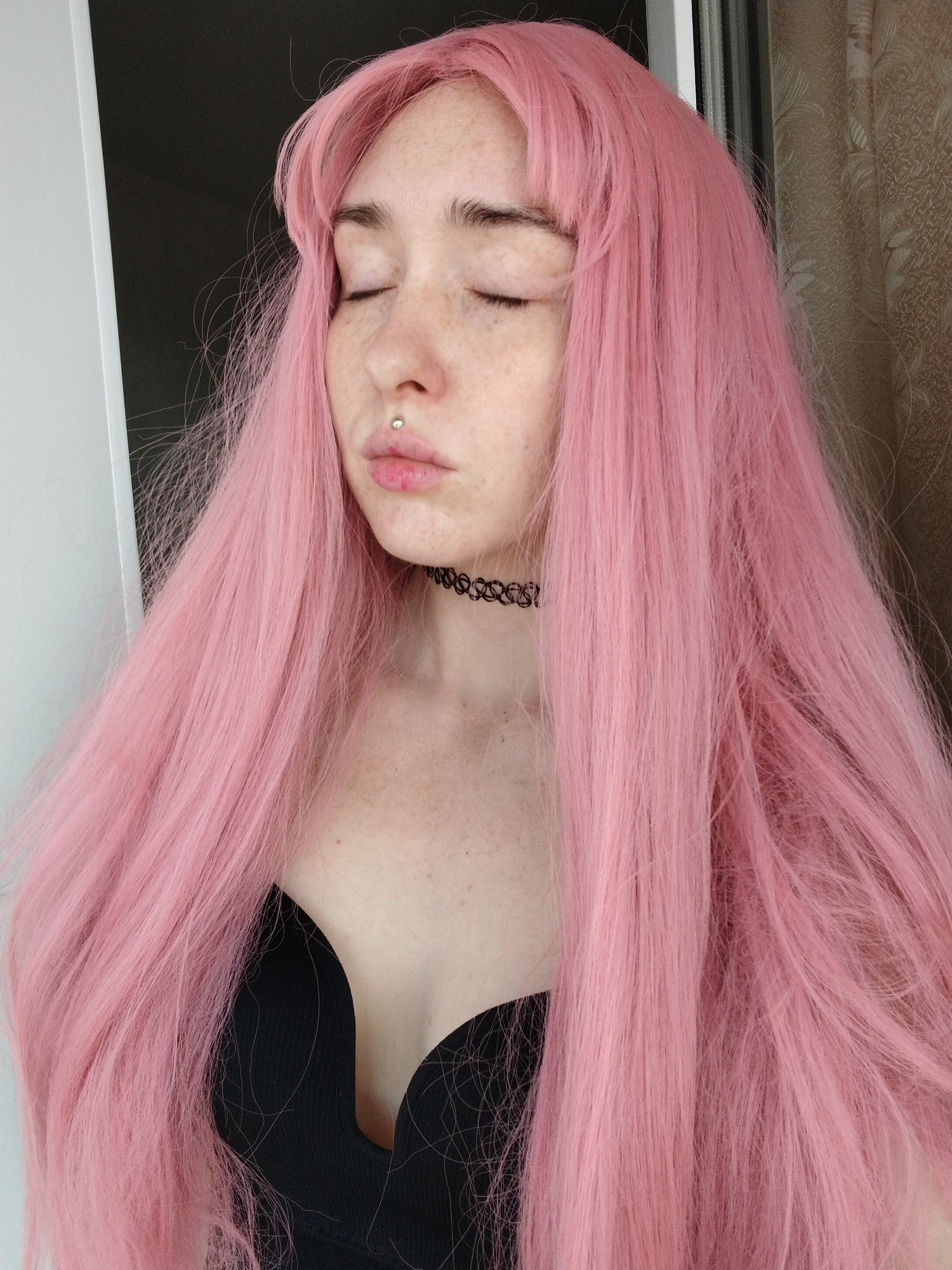 A young woman with closed eyes is centered in the frame. She has pale skin, freckles, and a lip piercing. Her hair is long, straight, and a light pink color. She wears a black, low-cut top and a black choker necklace. The background is a blurred mix of light and dark colors, possibly indicating an interior space. The image does not provide enough information to determine a specific location in Izhevsk, Russia.